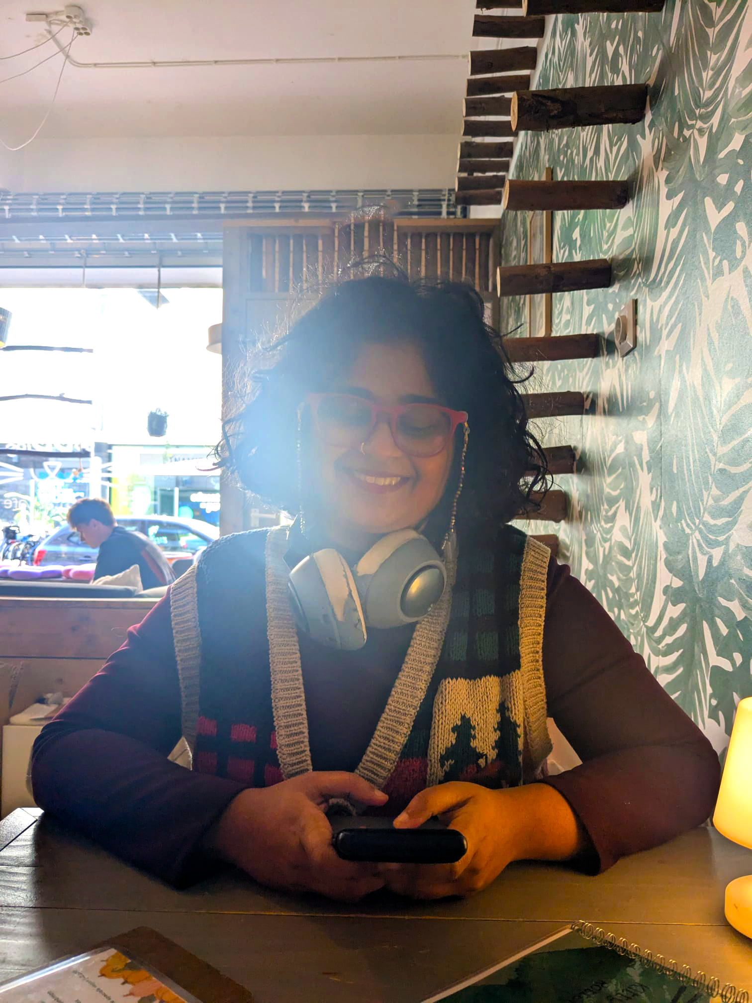 Woman with curly hair, red glasses, and headphones around her neck smiling while using a smartphone in a cafe or restaurant.