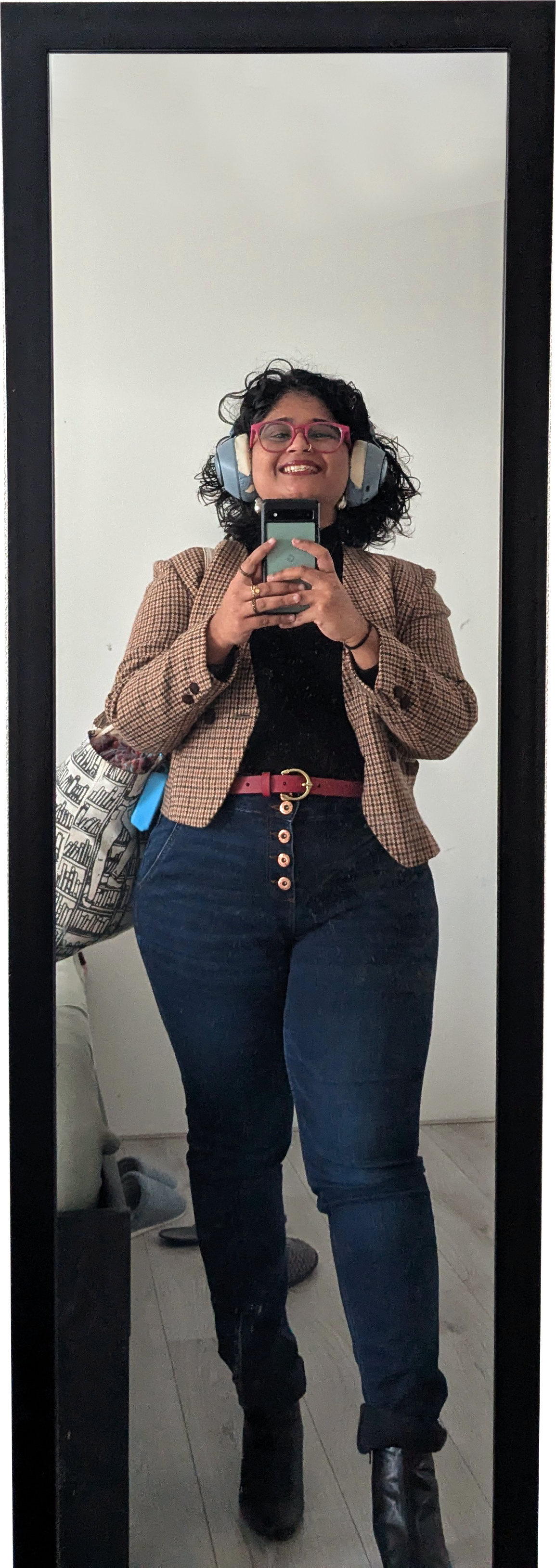 A woman taking a selfie in a mirror, wearing headphones, glasses, a checkered blazer, a black top, dark jeans, a red belt, black boots, and smiling.