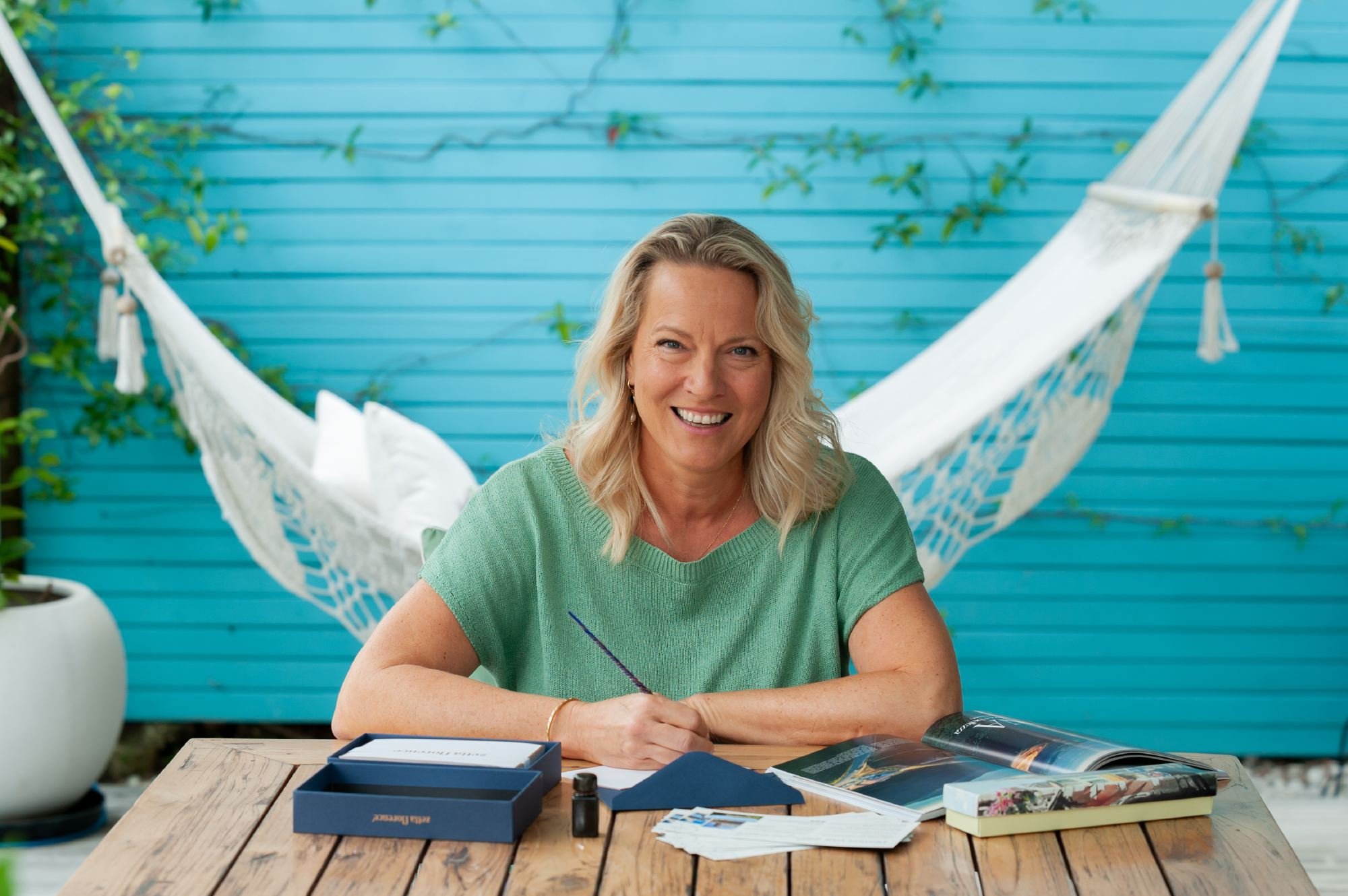 A woman with blonde curly hair smiling at a table outdoors in front of a blue wooden wall with vines, surrounded by various items including papers, a box, and a magazine.