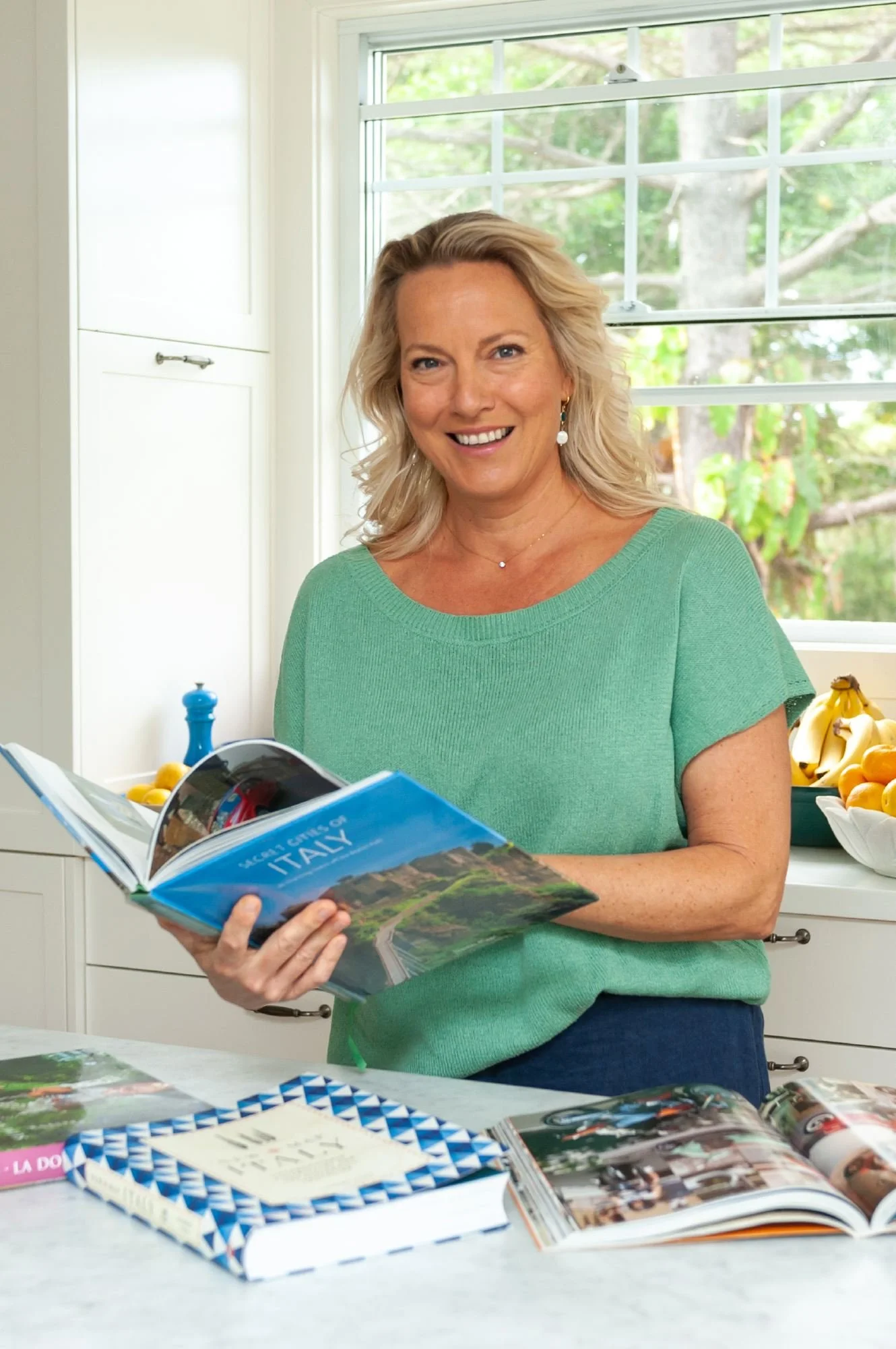 A woman with blonde hair smiling while looking at a travel magazine in a bright kitchen with a window showing trees outside.