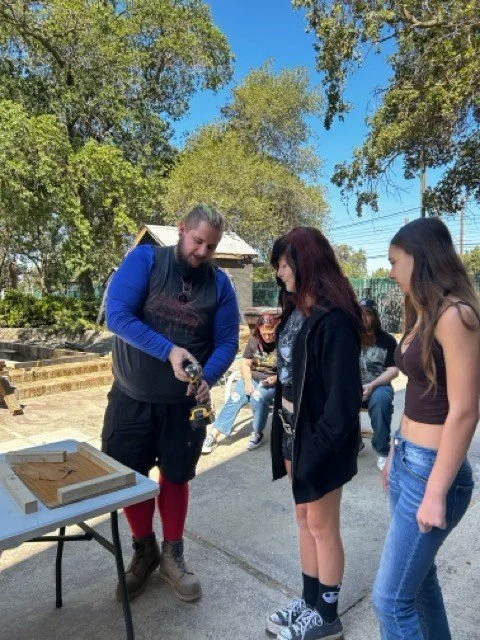 A man and two women standing outdoors, with the man demonstrating or showing something on his hand, while the women watch. There are trees and a small group of people sitting in the background.