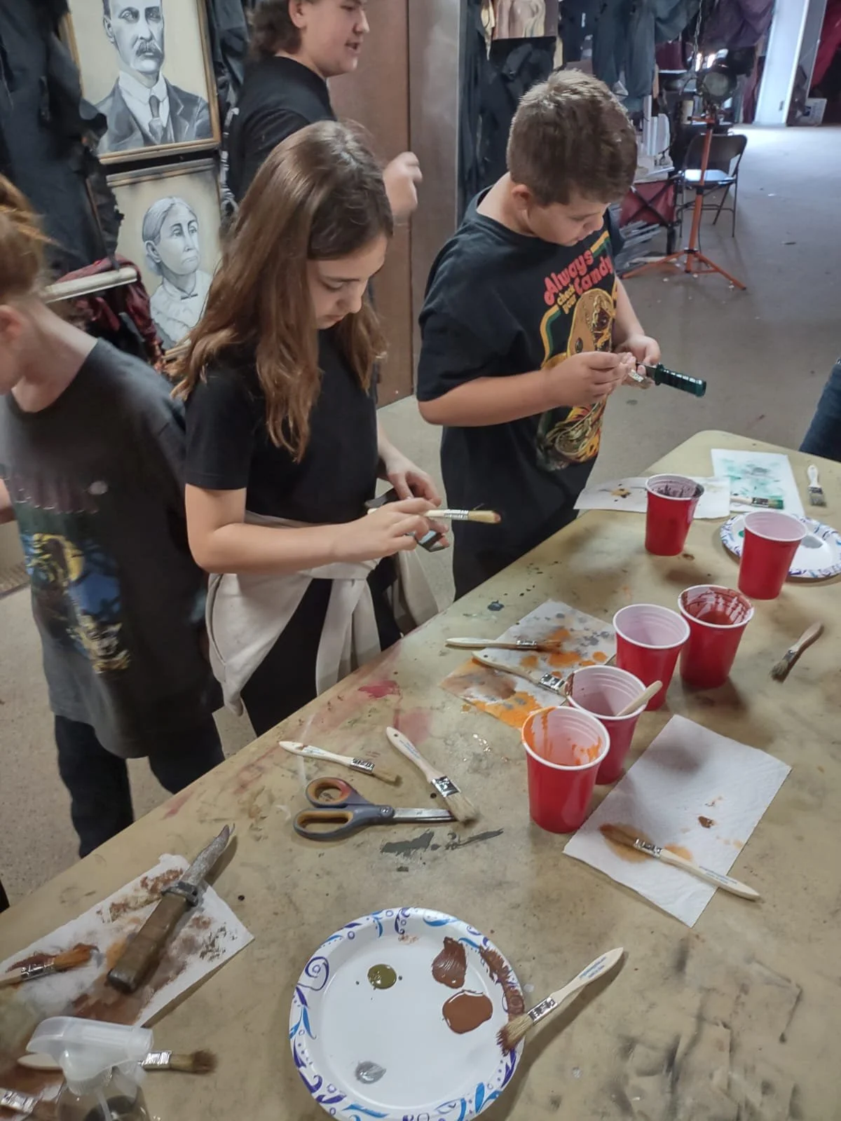 Children participating in a painting activity at a table with paint cups, brushes, and paper towels in an arts and crafts space.