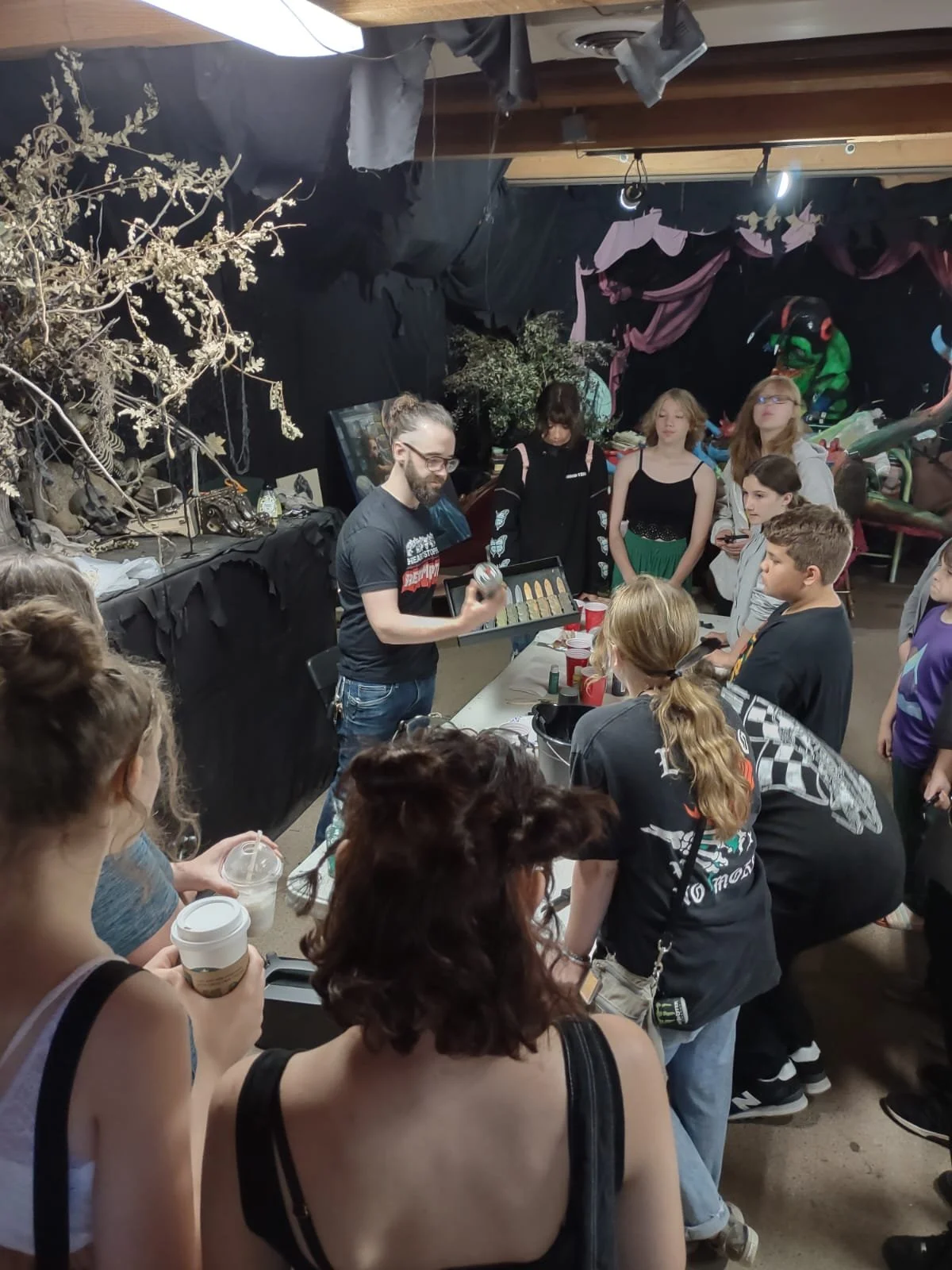 A man demonstrating with a spray can at a gathering of people in an indoor space decorated with black fabric, artificial trees, and props, while others watch.