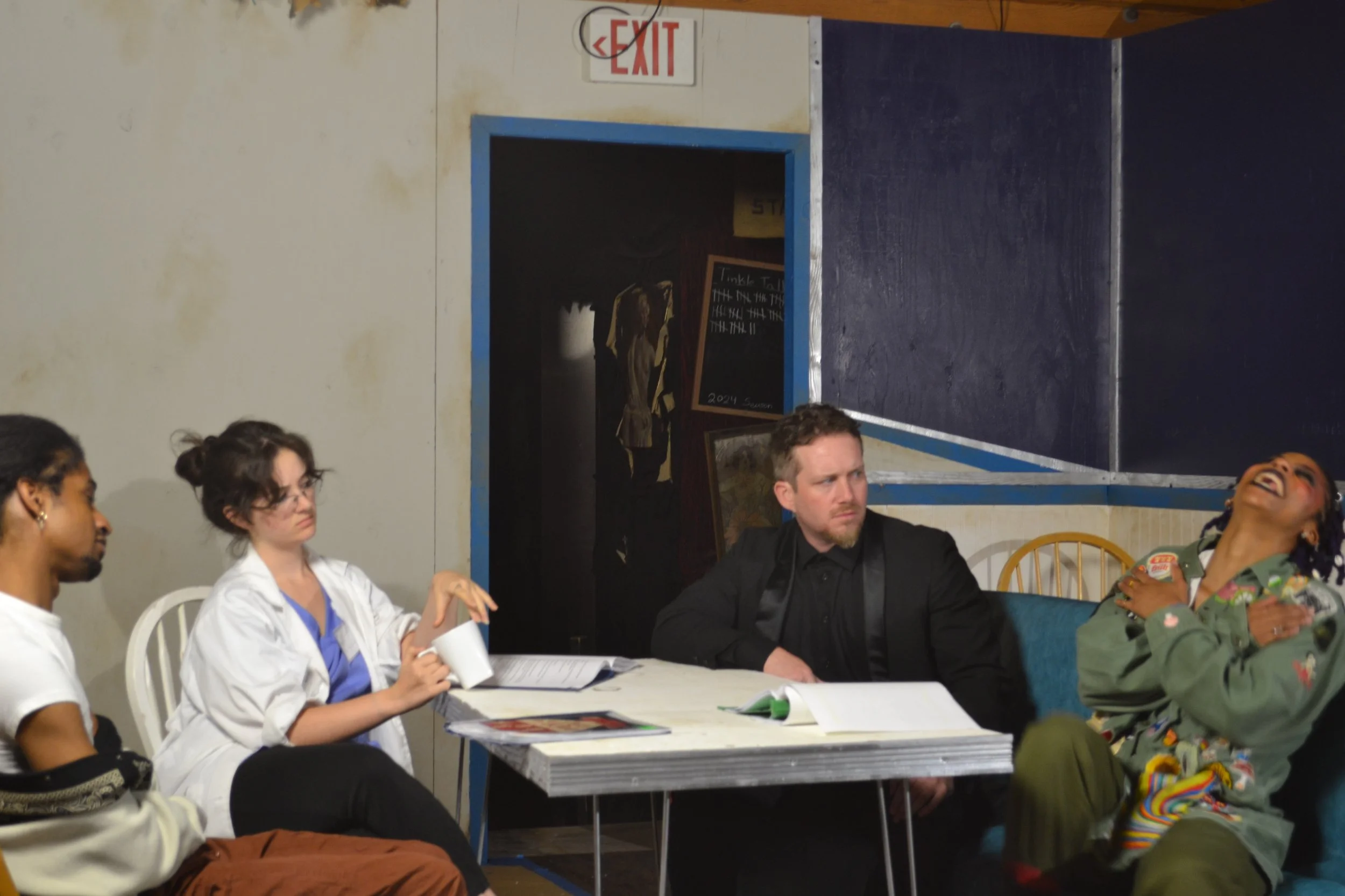 People sitting at a long table during a play rehearsal, with one person holding a cup and others listening attentively, in an indoor space with a visible exit sign and doorway in the background.
