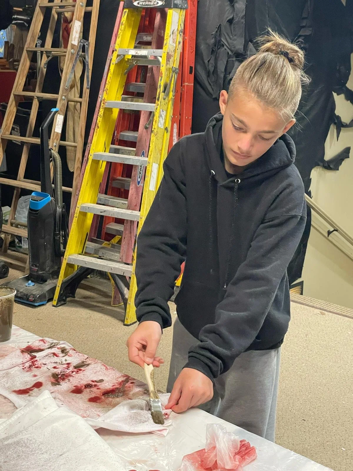 A young person with a ponytail, wearing a black hoodie and grey sweatpants, is preparing fish on a table covered with a white sheet with red bloodstains. There are ladders, a vacuum cleaner, and black backpacks in the background.