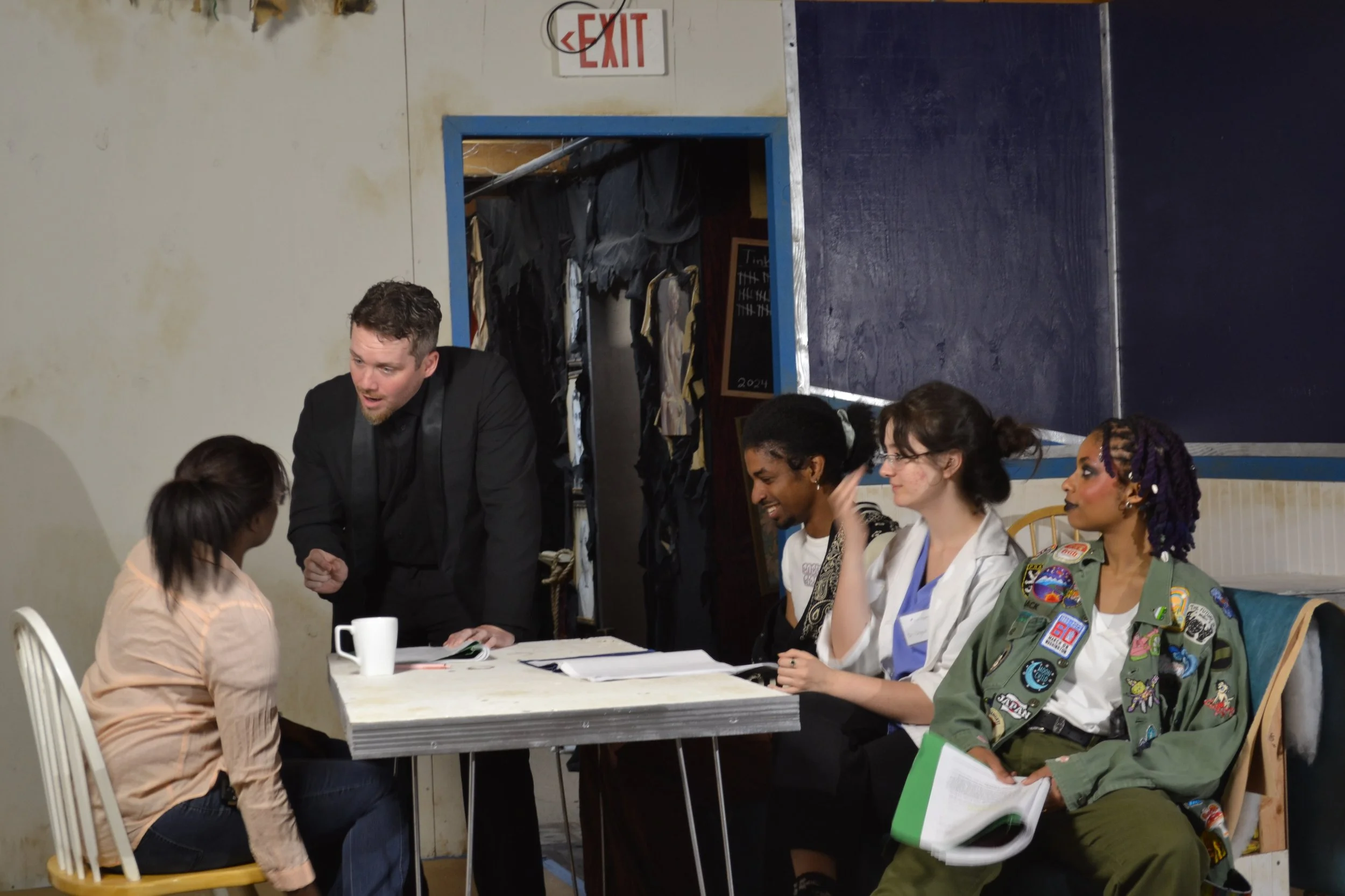 A group of diverse individuals inside a room engaged in conversation. Some are sitting at a table, and others are standing or sitting nearby. One person is dressed in a military-style uniform. There is an exit sign on the wall, and a doorway leading 
