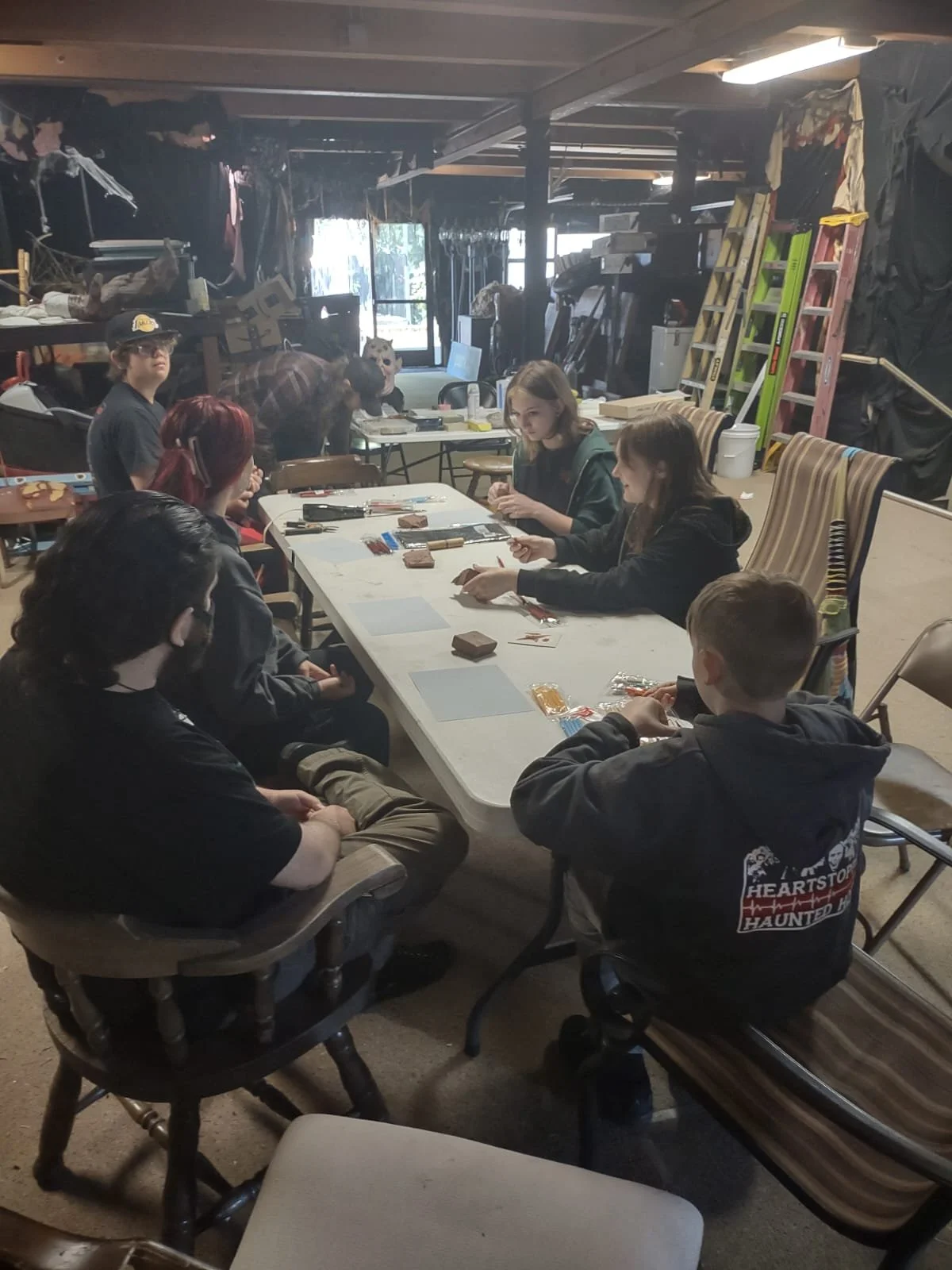 Group of people sitting around a table playing a card game inside a cluttered workshop or storage room