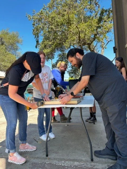 People gathered around a table outdoors, cutting or screwing wood together, with trees and a clear sky in the background.