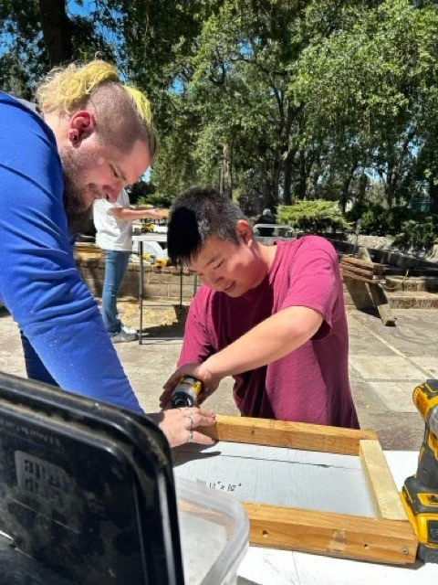 A man with long blond hair and a beard, wearing a blue hoodie, helping a young boy in a pink shirt on a woodworking project outdoors. The boy is using a drill on a wooden frame while the man supports him. There are trees and other people in the backg