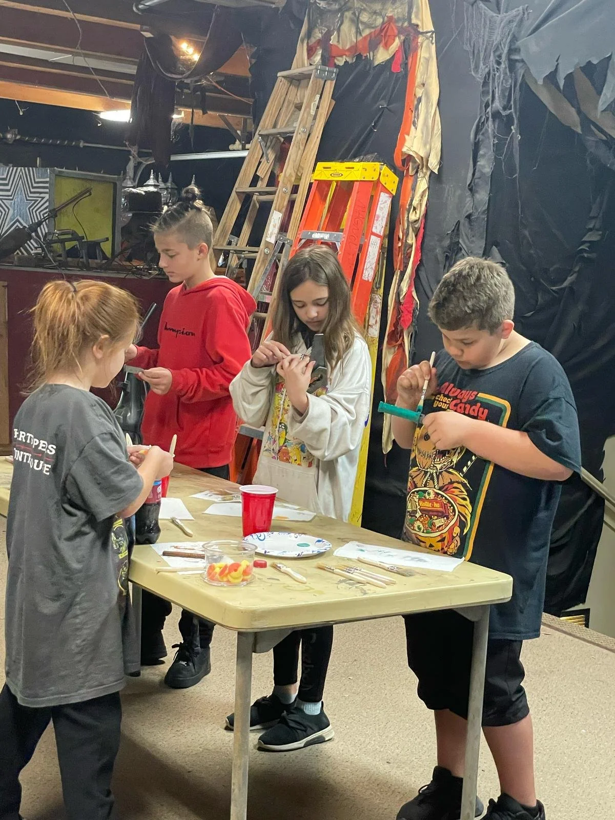 Four children standing around a table covered with art supplies, working on crafts in a room with ladders and black plastic sheets in the background.