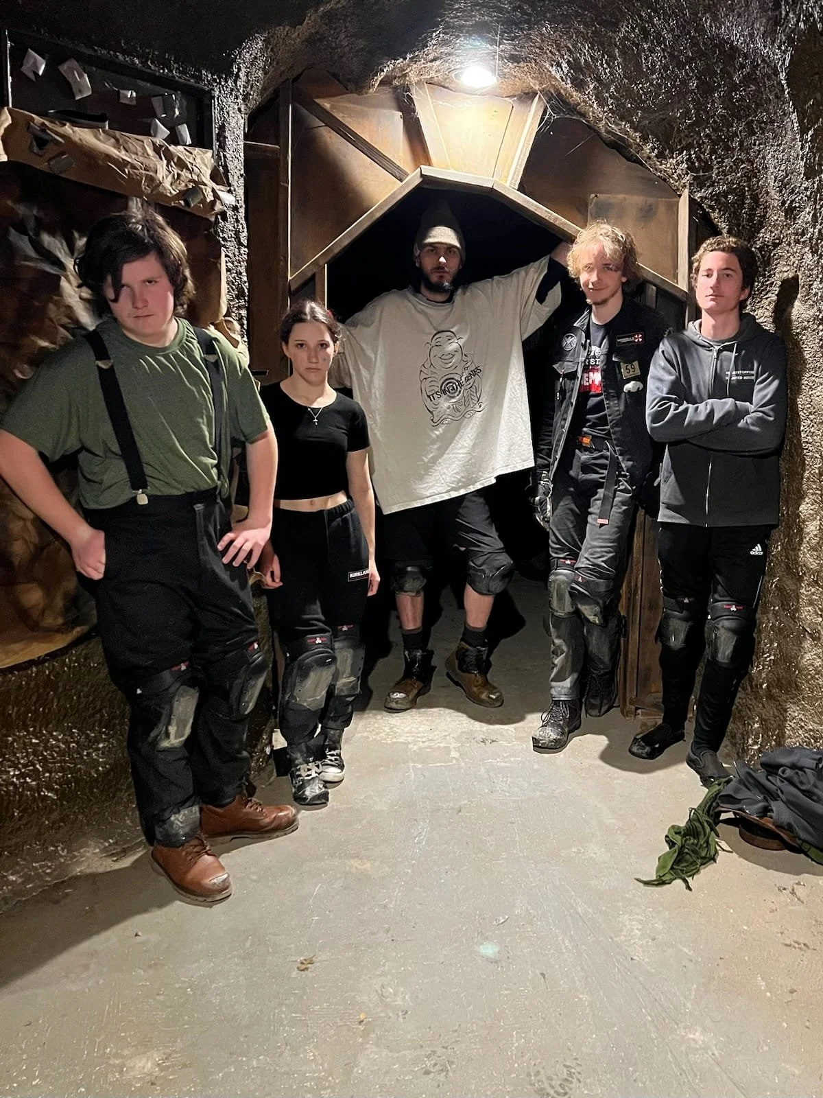 Five people standing inside a tunnel or underground shelter, dressed in casual and outdoor clothing, with some wearing knee pads and boots, posing for the photo.