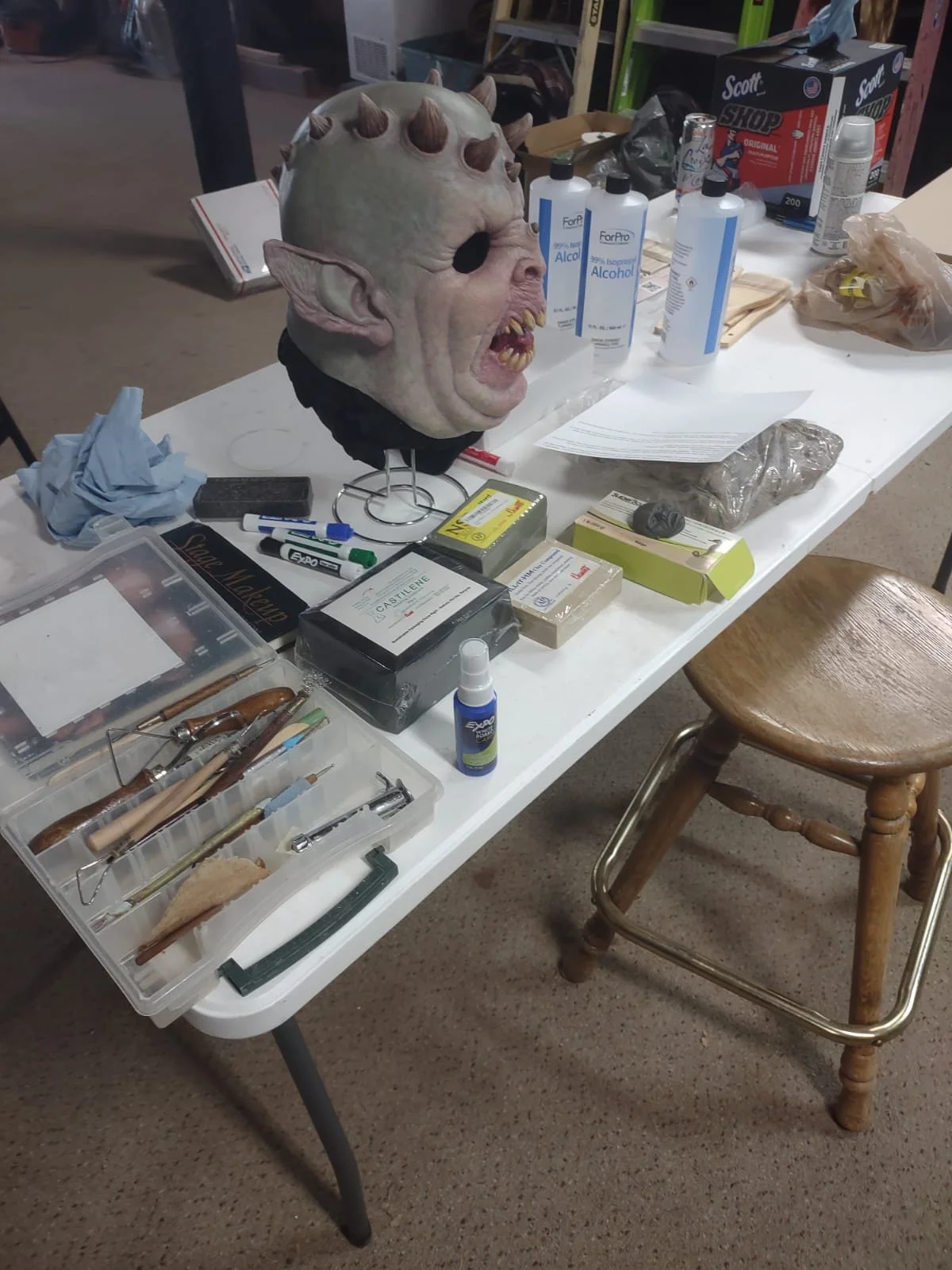 A creepy, creepy Halloween mask of a monstrous face with horns, large ears, and sharp teeth sits on a table surrounded by tool kits, bottles of alcohol sanitizer, and various paper and boxes, with a wooden chair nearby.