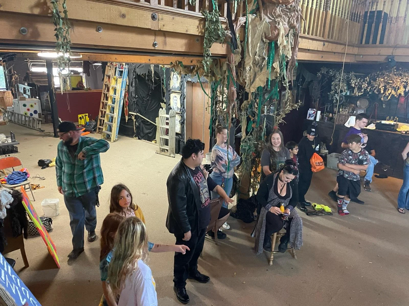 People gathered in a rustic indoor space with wooden walls and a high ceiling, some sitting and others standing, surrounded by hanging vines and decorative foliage, with various items and artwork visible in the background.