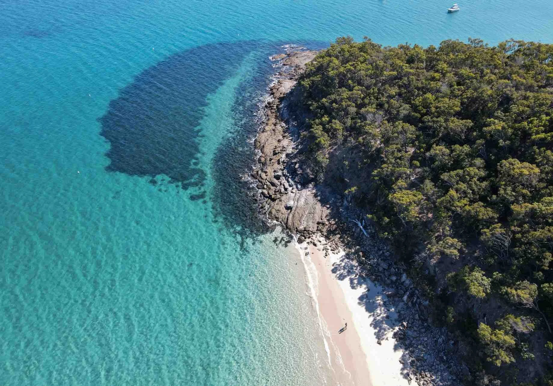Aerial view of a small beach with turquoise water, rocks, and dense green forested shoreline, with boats anchored nearby.