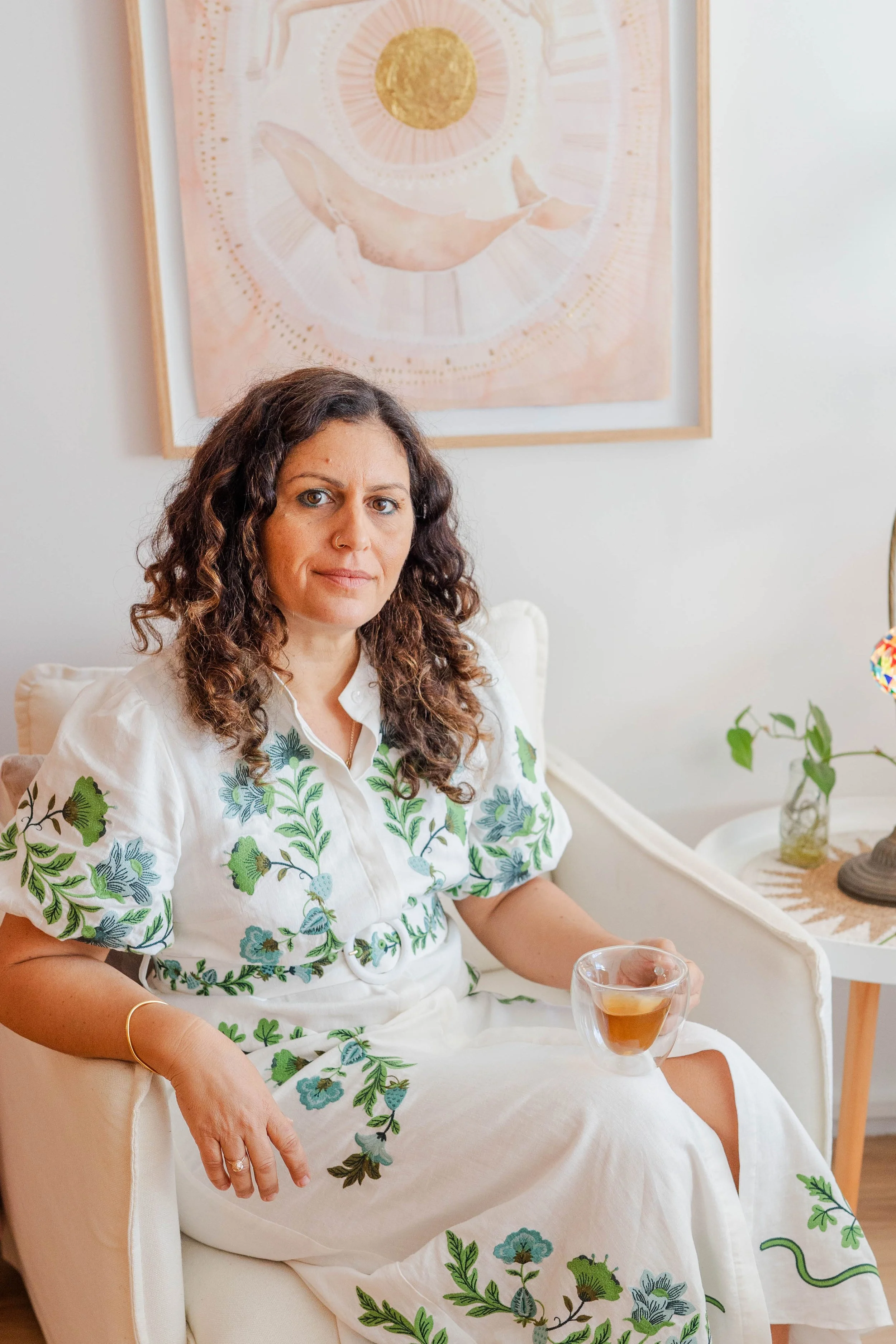 A woman with curly brown hair sitting on a cream-colored armchair in a well-lit room. She is wearing a white dress with green and blue floral embroidery and holding a glass cup filled with a dark beverage. There is a framed artwork behind her depicting a whale, the sun, and moon in pastel colors. A small side table with a potted plant and a decorative lamp is nearby.