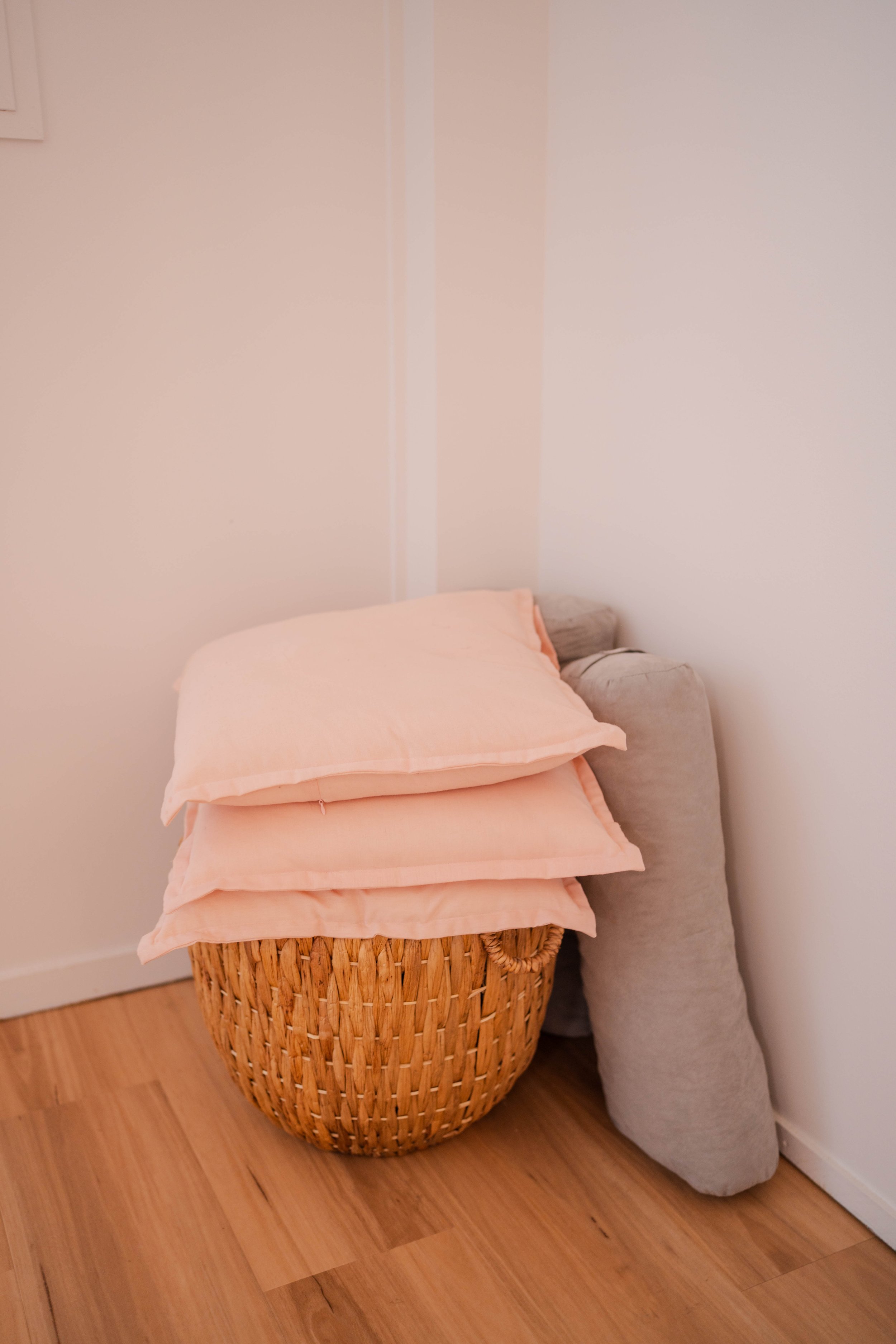A wicker laundry basket with three folded pink bedsheets on top, leaning against a beige cushion, in a corner of a room with wooden floors and white walls.