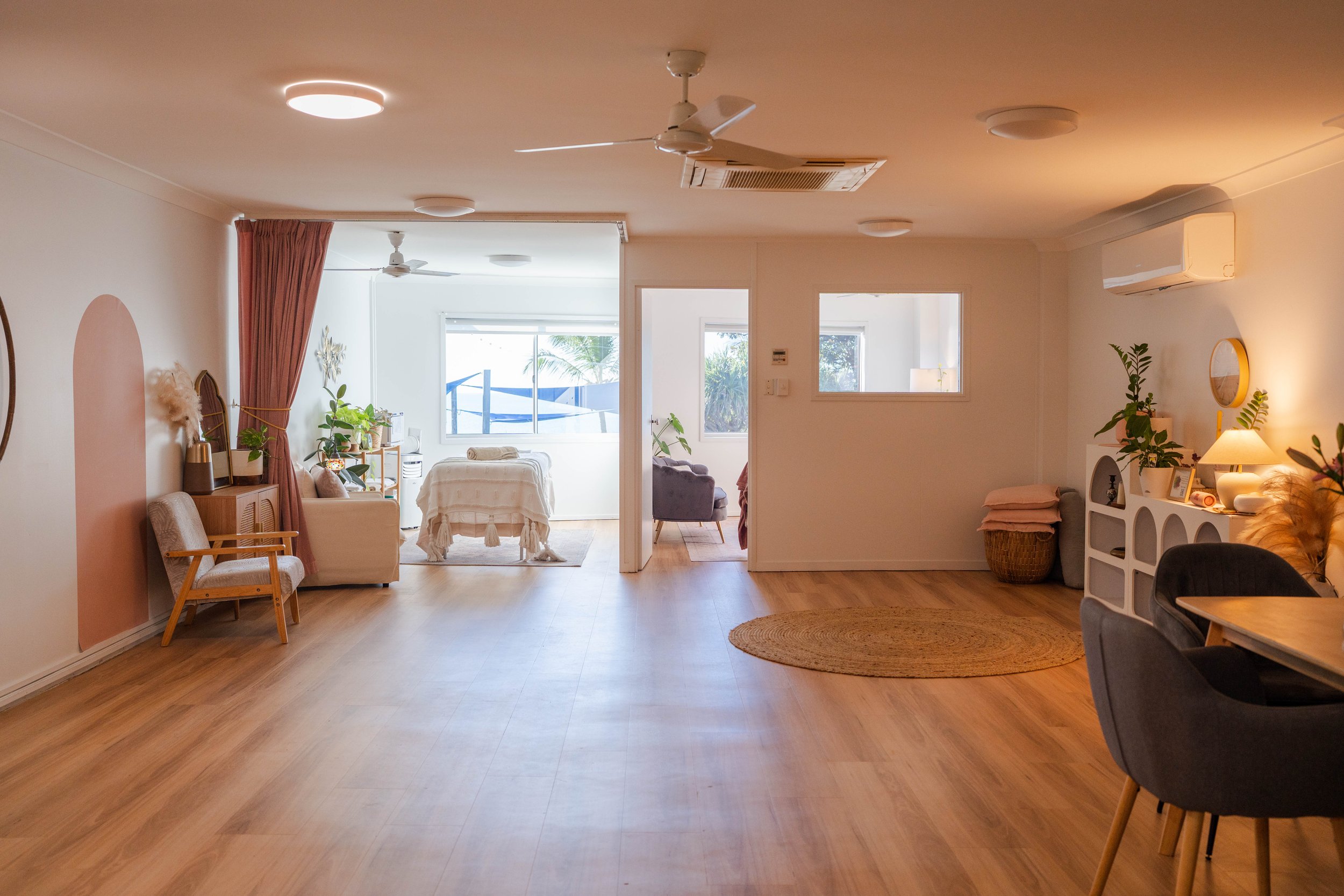 Spacious living room with wooden flooring, white walls, and modern decor. The room has a ceiling fan, air conditioning unit, and multiple light fixtures. There are various plants, a round woven rug, a white shelf with framed photos and decorative items, and seating areas including a chair, sofa, and armchairs. Large windows let in natural light, and there is a view of an outdoor space with trees and palm leaves.