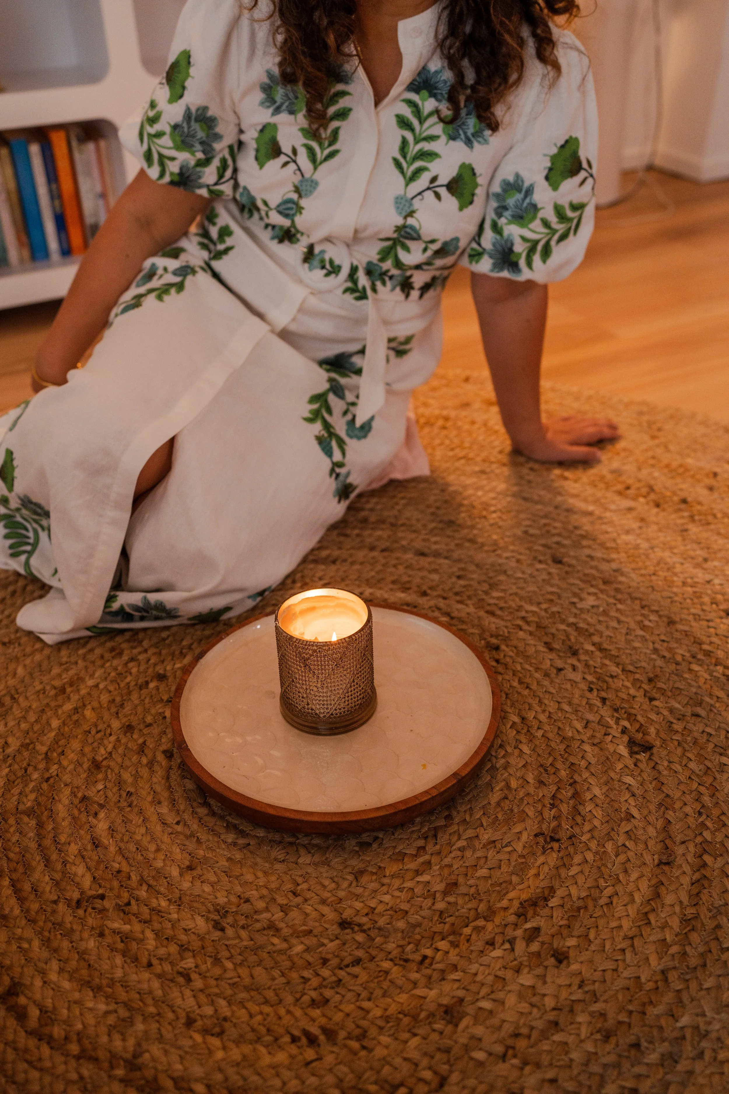 Person kneeling on a woven rug, dressed in a white dress with green and blue floral patterns, next to a lit candle on a circular wooden tray.