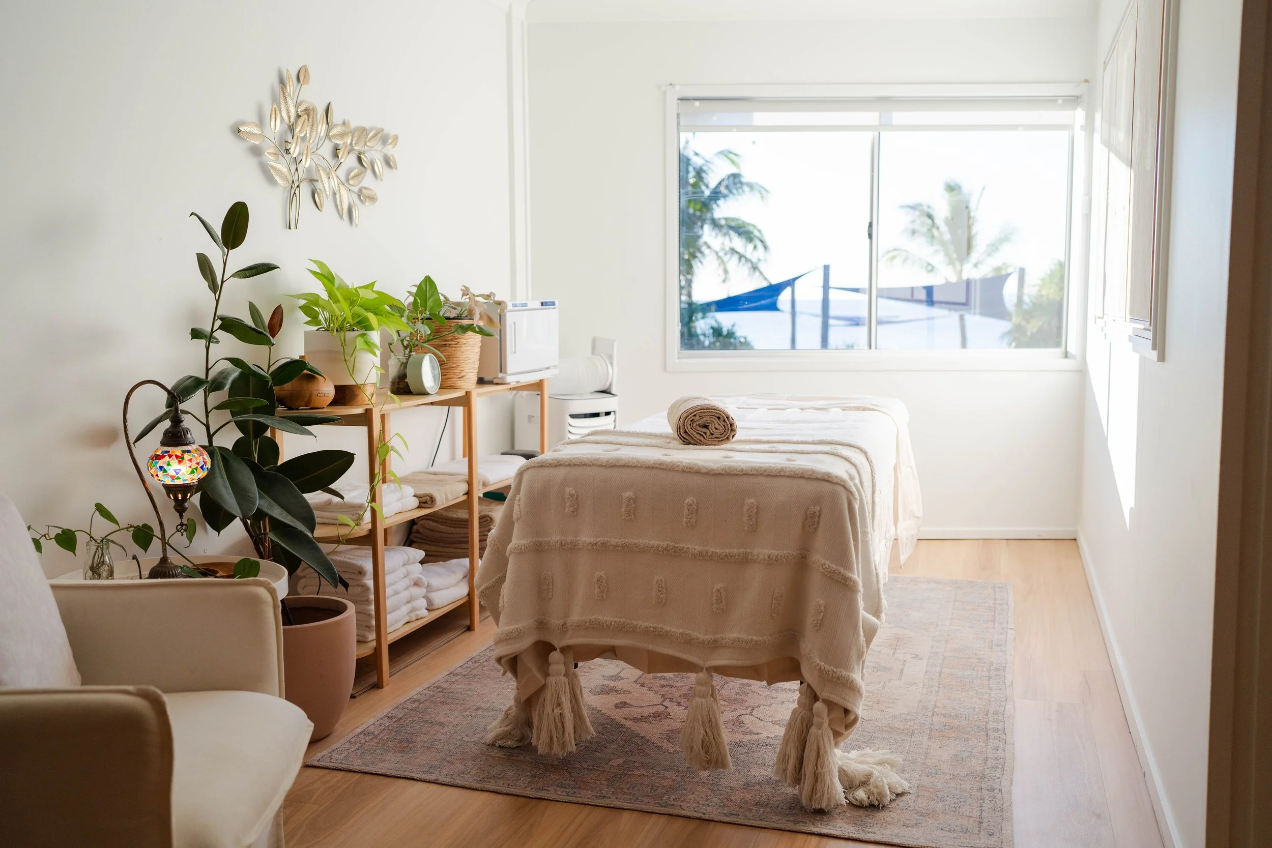 A massage room with a massage table covered in a beige towel, a rolled towel on top, and natural light coming from a large window with a view of palm trees outside.