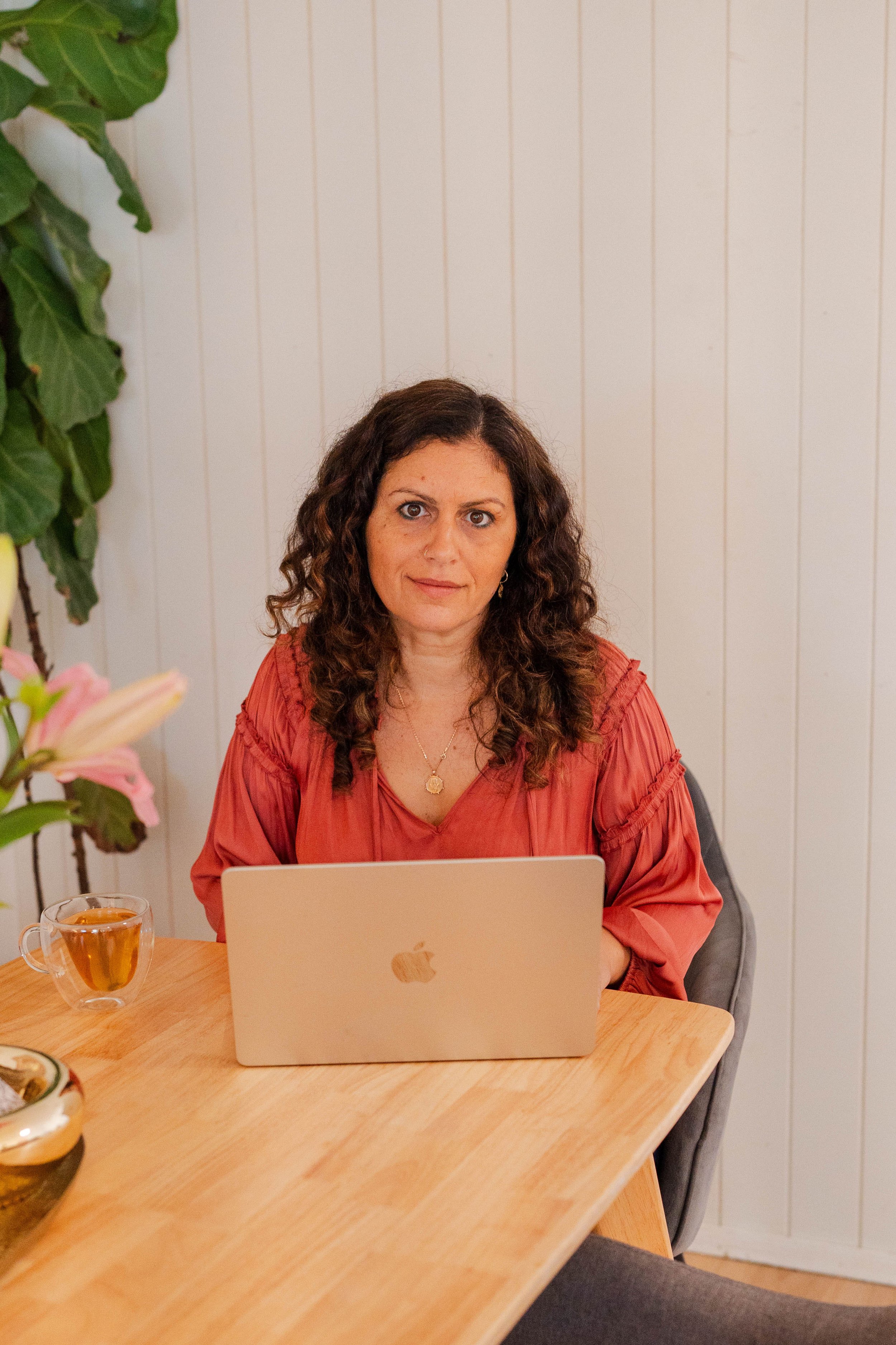 A woman with curly brown hair and light skin, sitting at a wooden table with an open laptop in front of her, wearing a coral-colored blouse, in a room with white wall paneling and a plant to her left. There is a glass cup with tea and some flowers on the table.