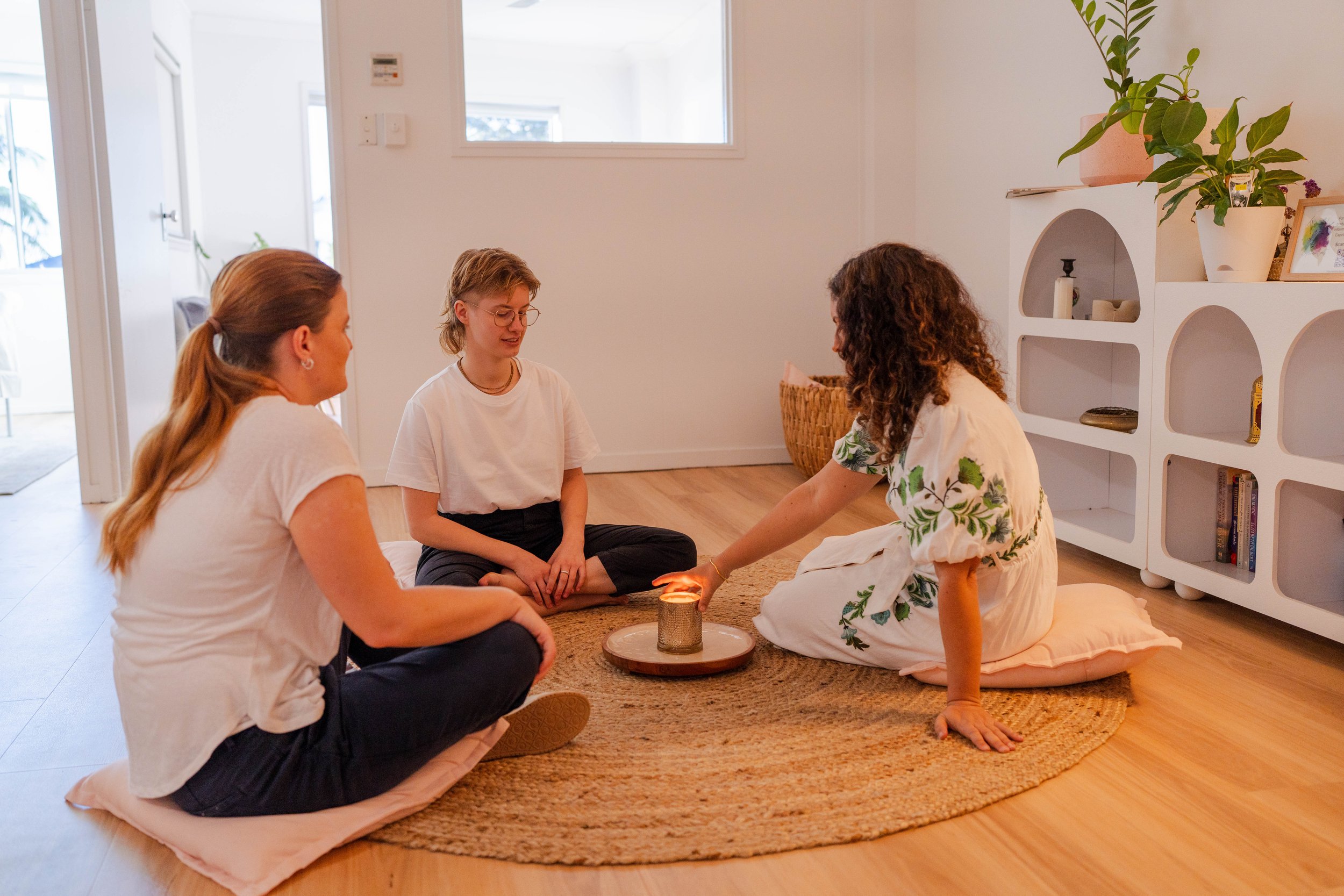 Three women sitting on a round woven rug indoors, participating in a ceremony involving a candle box with flame, in a cozy, well-lit room with white walls, a bookshelf, and plants.