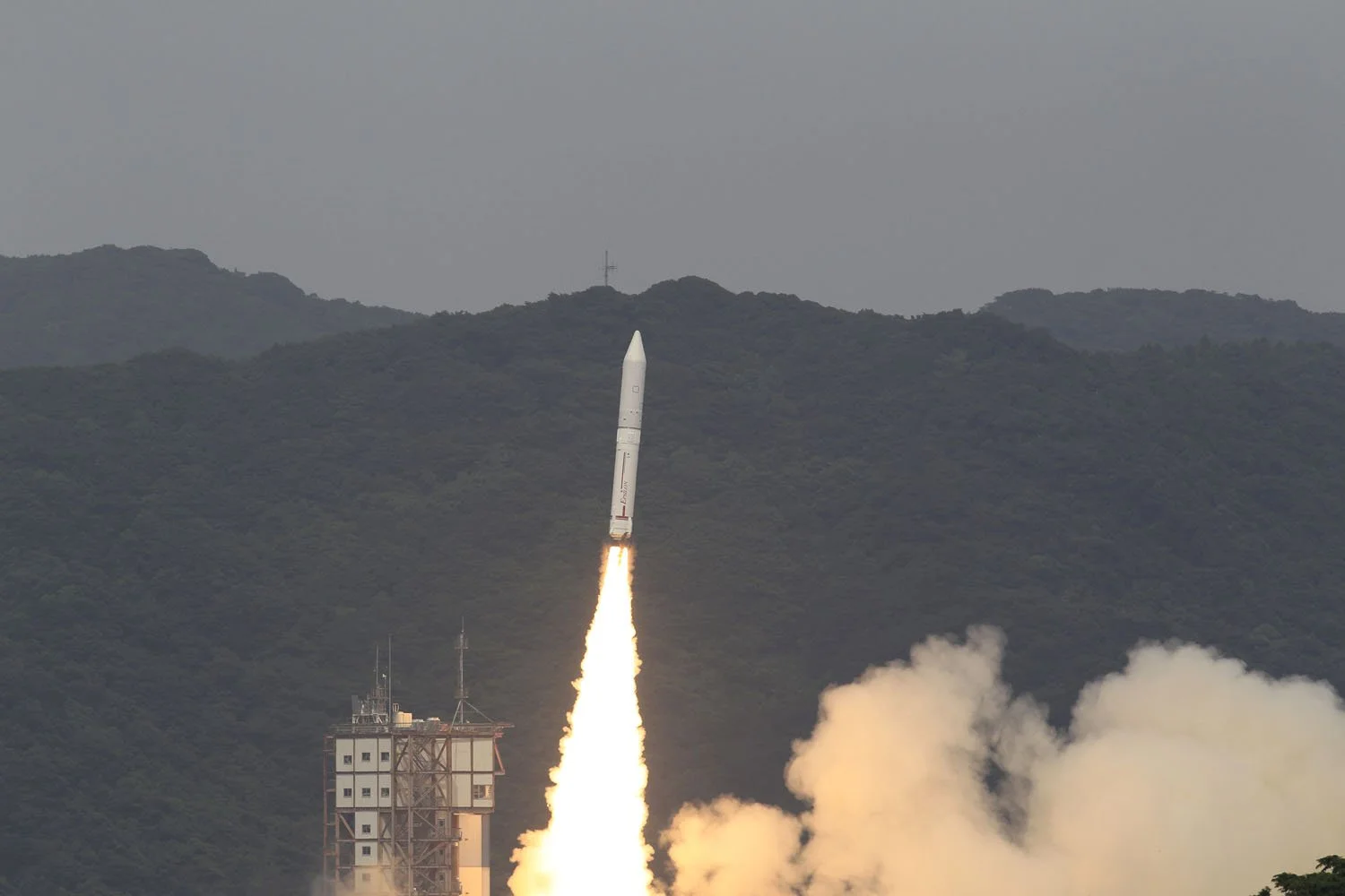 Epsilon launching from a tall building with a mountain range in the background, smoke and flames visible at the base of the rocket.