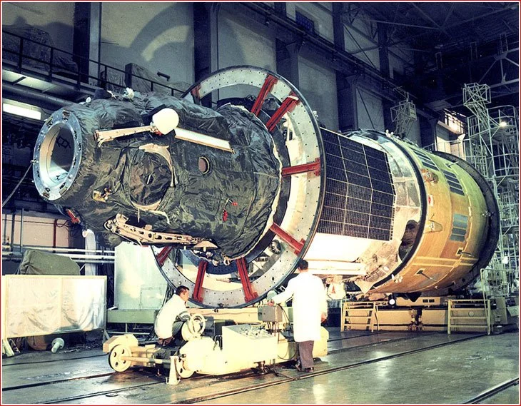 Engineers or scientists working on a large spacecraft or satellite in a manufacturing or assembly facility.