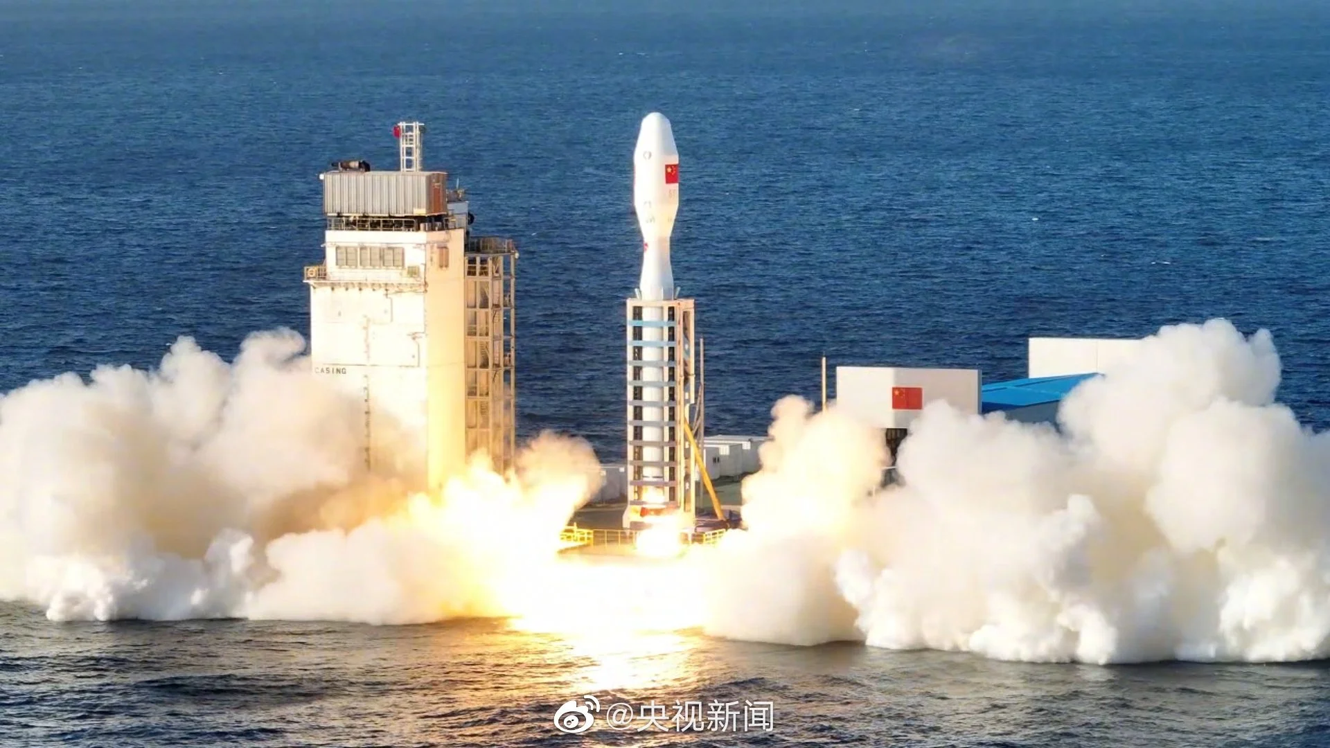 A rocket on a launch pad during takeoff, with smoke and fire at the base, ocean in background, Chinese flags visible on the rocket and nearby structures.