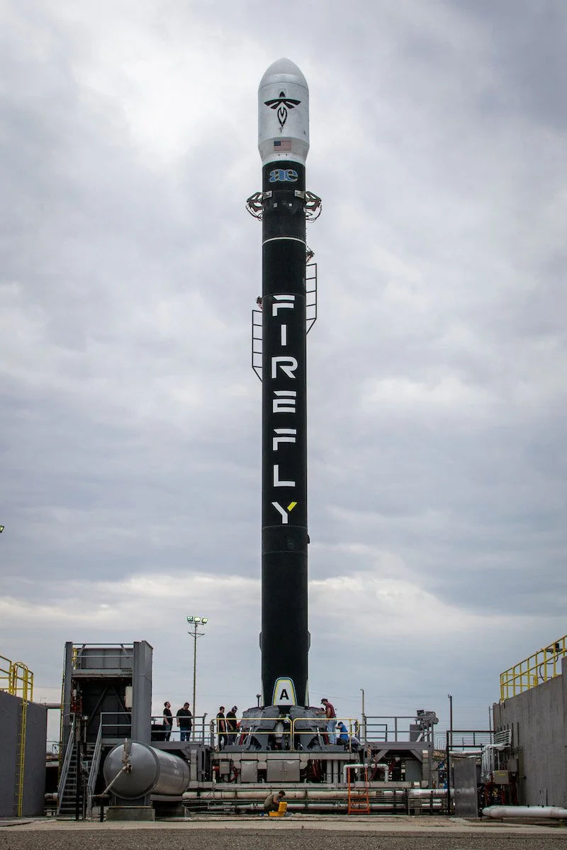 Alpha stands vertically on a launch pad against a cloudy sky, with several people working around it.