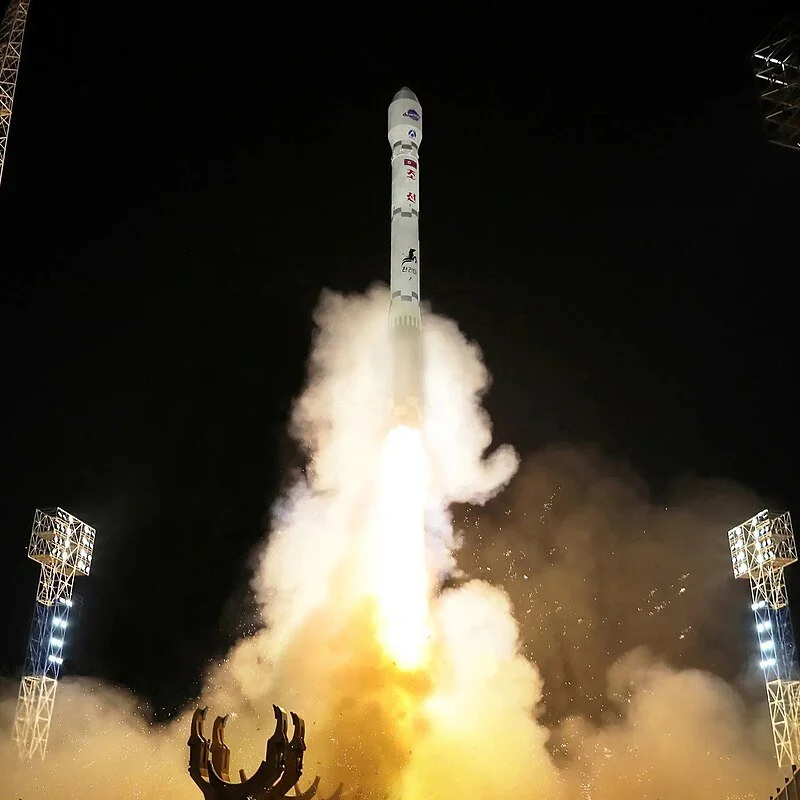 Chollima-1 launching at night with flames and smoke, illuminated by bright flares and surrounded by structures.