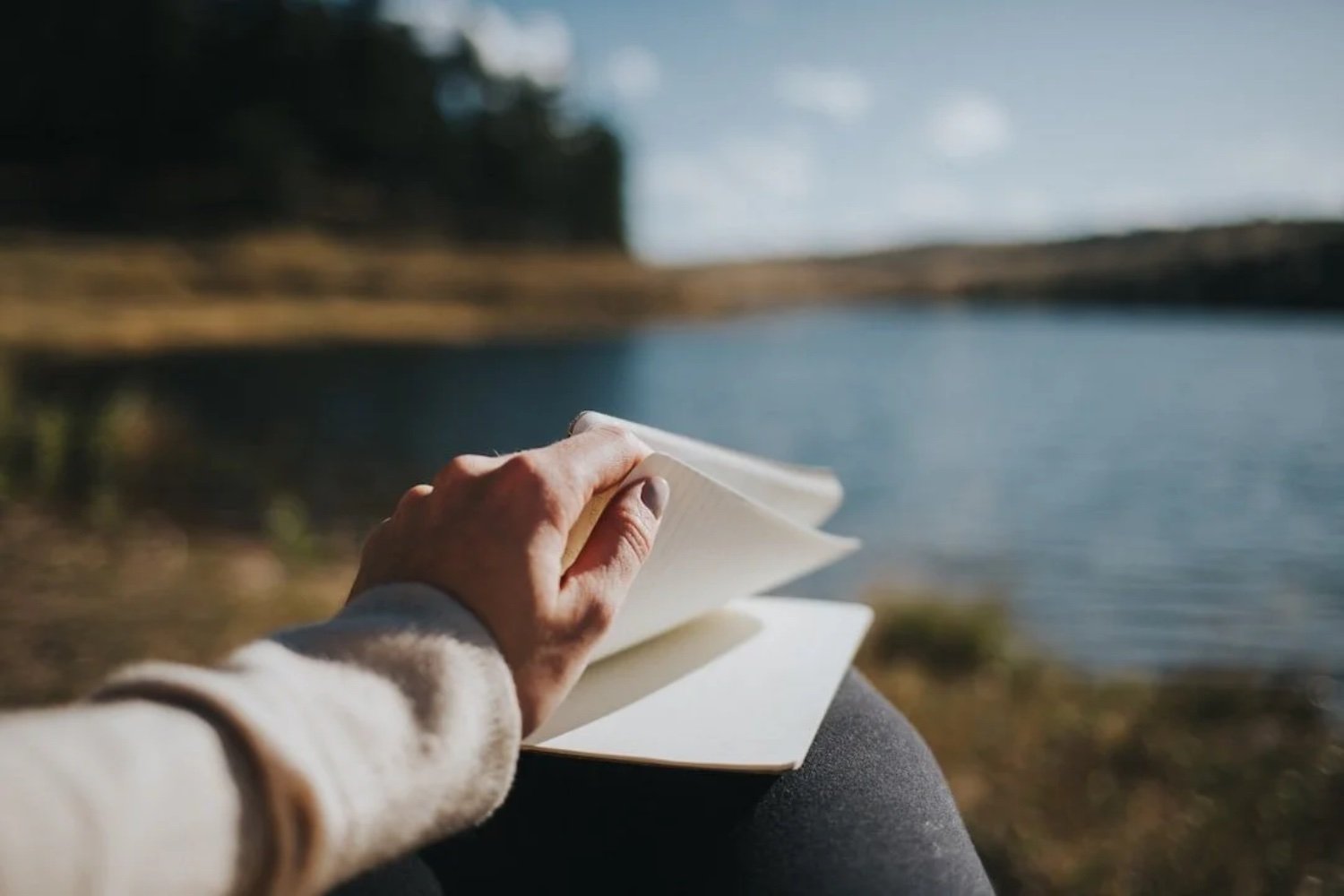 Person holding an open notebook near a body of water with trees in the background on a clear day.