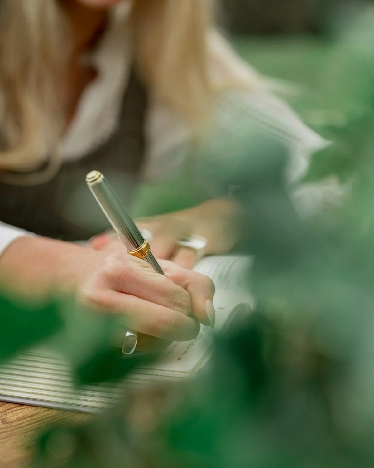 Close-up of a person's hand holding a silver pen, writing in a lined notebook, with blurred green foliage in the foreground.