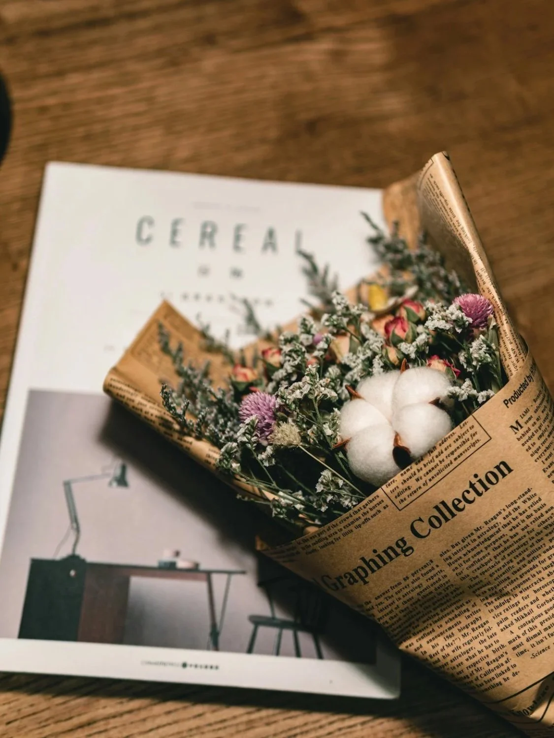 A bouquet of flowers wrapped in vintage newspaper rests on a magazine titled 'Gereat' on a wooden surface.