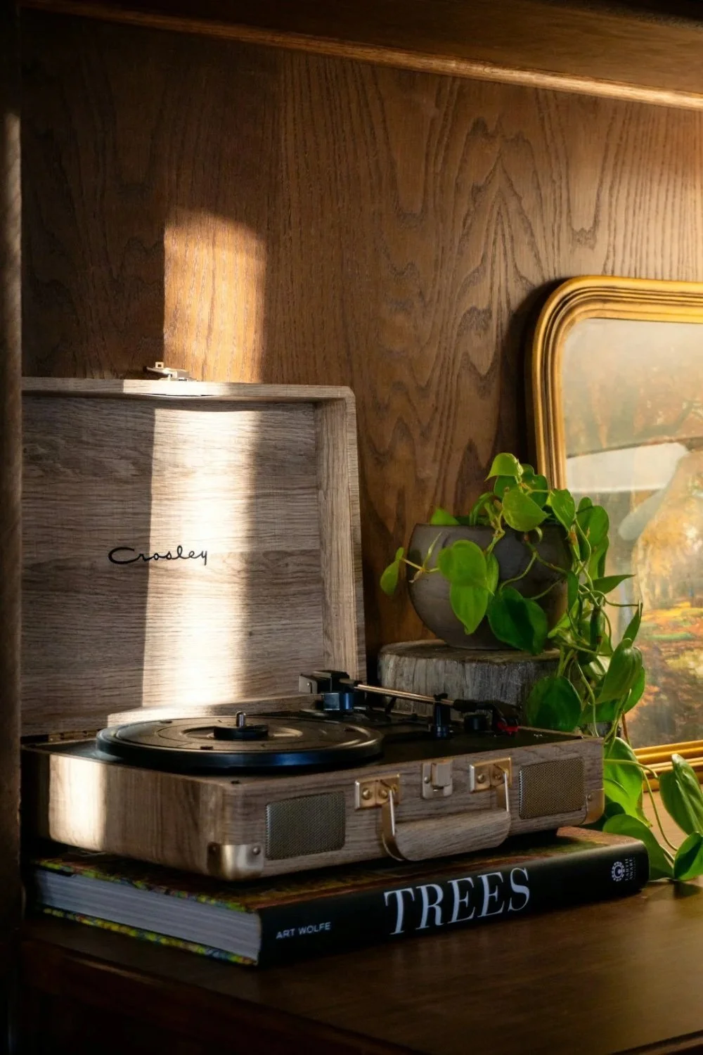 A vintage Crosley turntable in a wooden case sitting on top of a large book titled 'TREES' by Art Wolfe, with a potted green plant and a framed picture in the background.