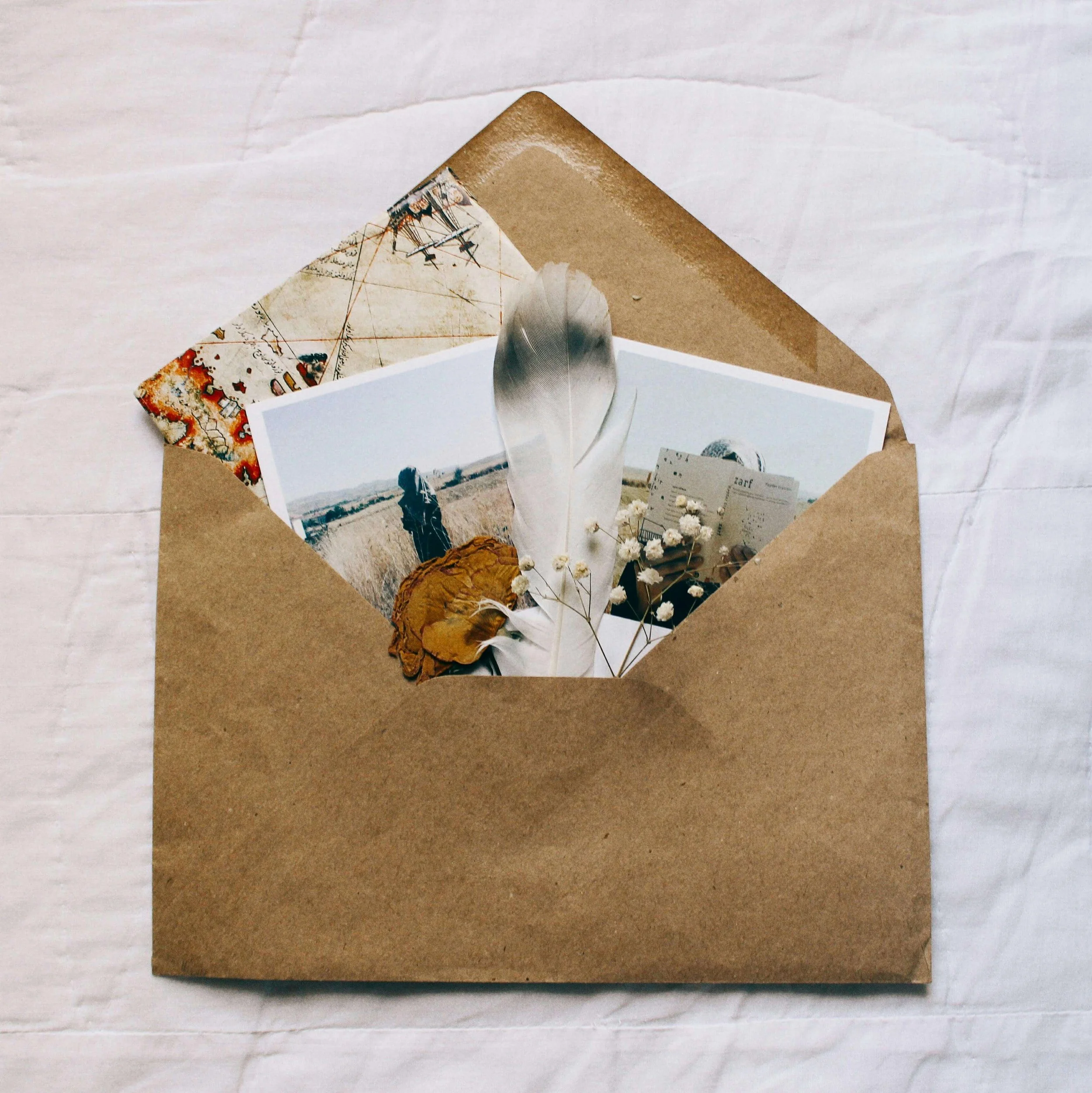 Open brown paper envelope containing photos, dried flowers, a large feather, and small white flowers, placed on a light fabric surface.