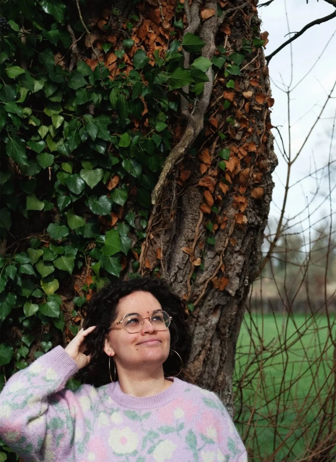 A woman with curly black hair, glasses, and hoop earrings smiles while touching her head, standing outdoors next to a large tree with green ivy and brown leaves. In the background, there is a field and leafless trees.