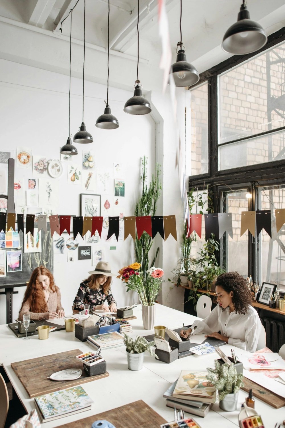 Three women sitting at a large white table in a bright, decorated art studio. They are drawing or working on creative projects. The room has plants, framed pictures on the wall, and a row of black and red triangular banners hanging across the window. There are vases of flowers on the table and art supplies around.