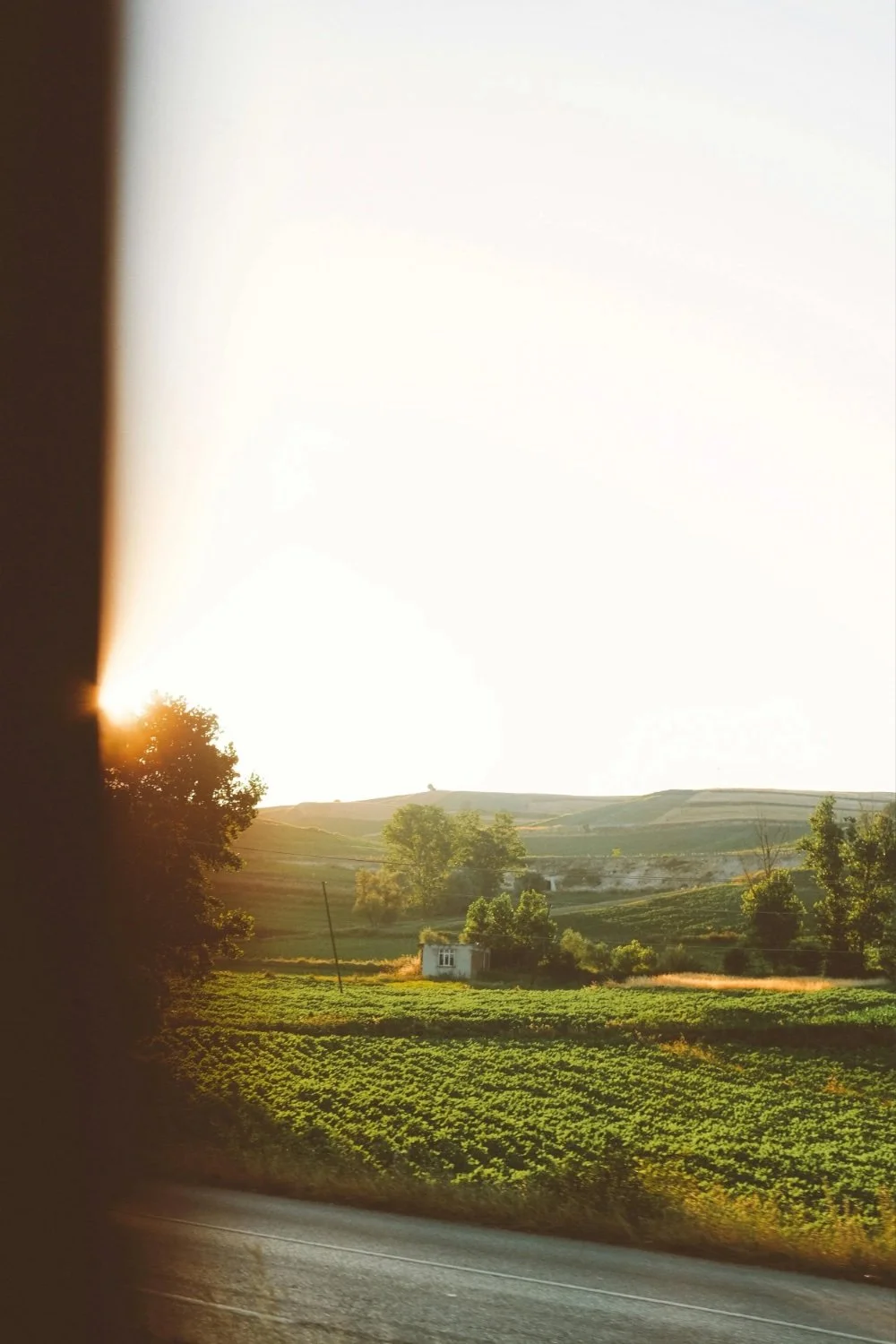 Sunset over a lush countryside landscape with trees, green fields, and a small house, viewed through a window with shadowed edges.