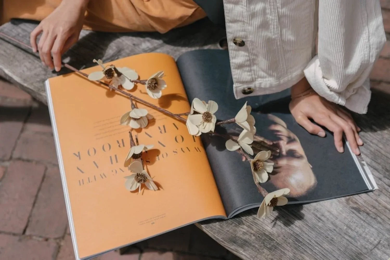 Open magazine with a branch of cream-colored flowers placed on it, on a rustic wooden table with a person in a light-colored jacket and orange shirt leaning over.