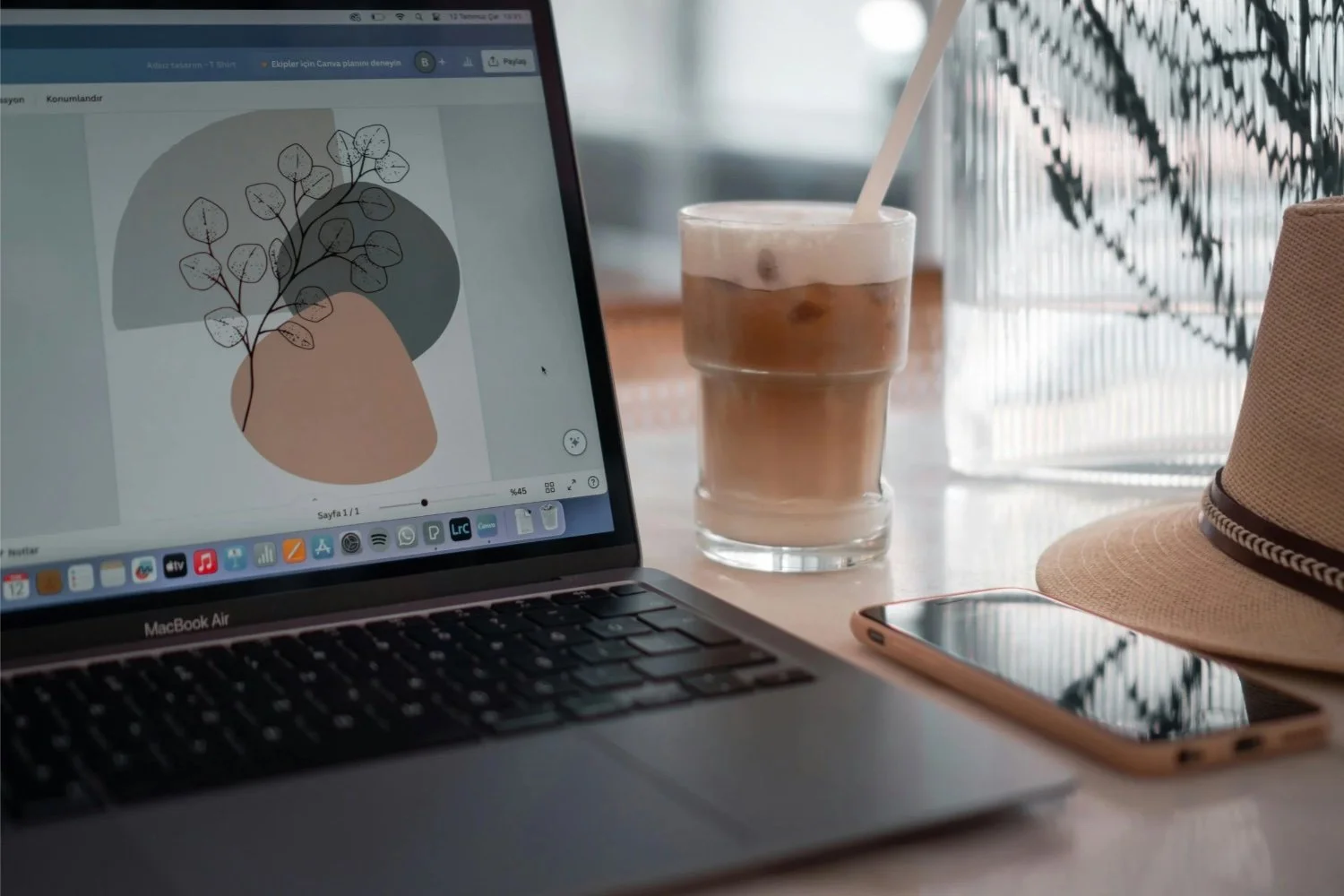 Laptop on a desk displaying a design project, next to a glass of iced coffee with a straw, a smartphone, and a beige hat with a black band, with a window and a plant in the background.