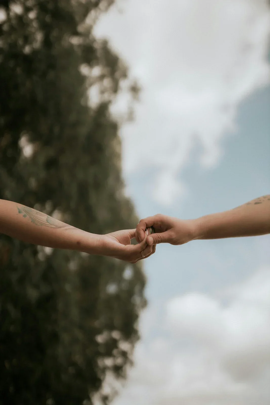 Two people holding hands outdoors, with a blurred landscape and sky in the background.