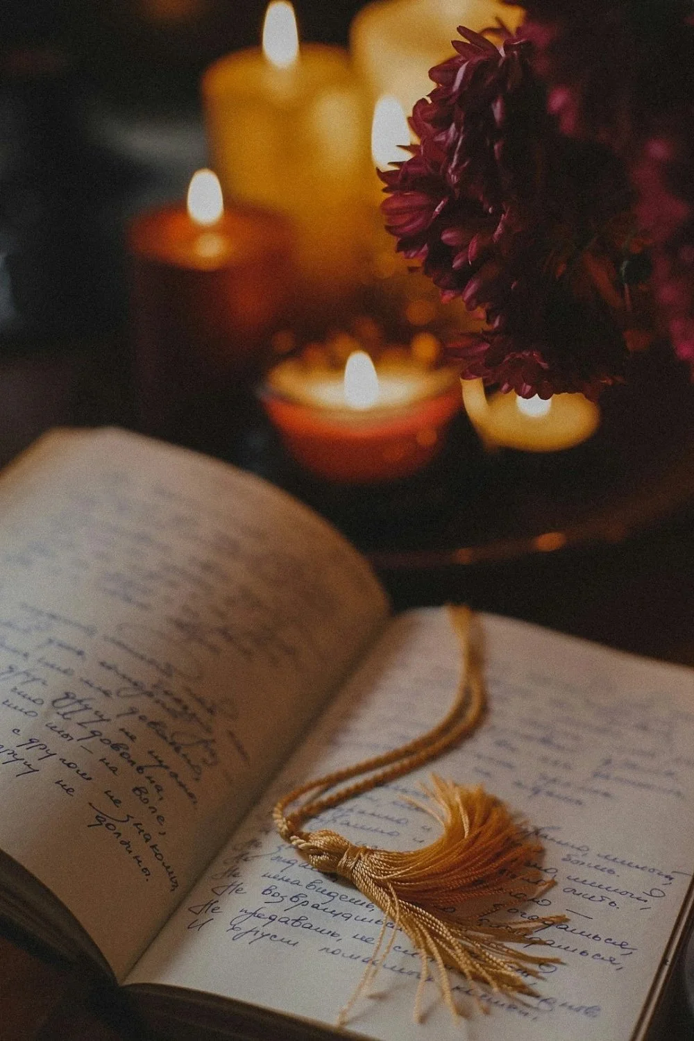 An open handwritten journal with a tassel bookmark in the foreground, lit candles and a dark purple flower in the background creating a warm, cozy atmosphere.