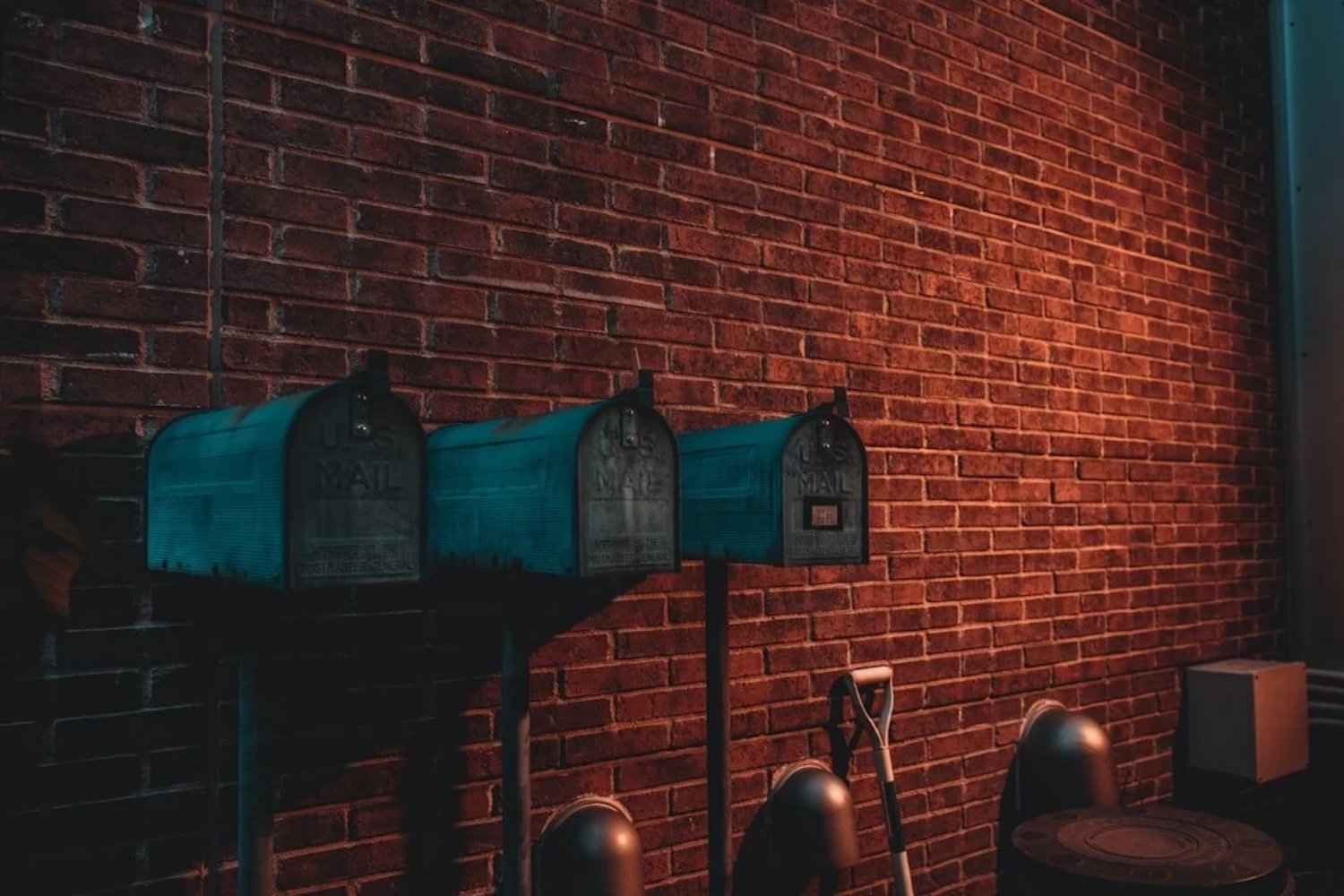 Four blue USPS mailbox units mounted on a black stand against a red brick wall, with a shovel, a metal object, and a white box beneath them.