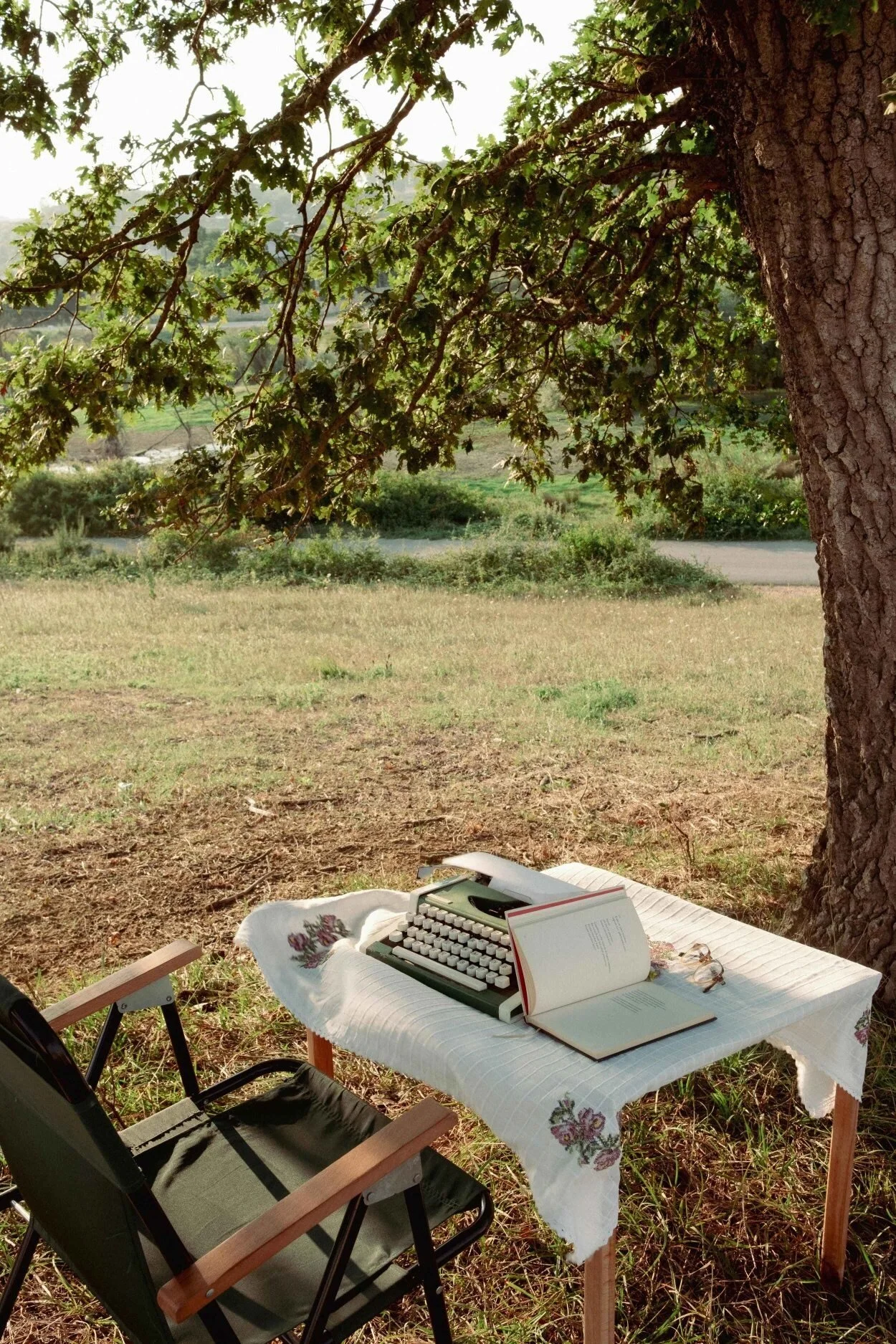 A table with a quilted cloth, a vintage typewriter, and an open book, set outdoors under a large tree with greenery in the background.