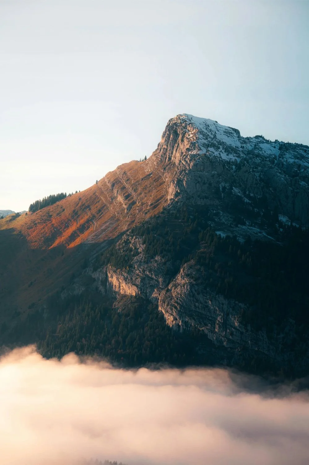 A mountain with snow-capped peak, rocky slopes, and patches of trees, surrounded by clouds or fog at the base, during daytime.