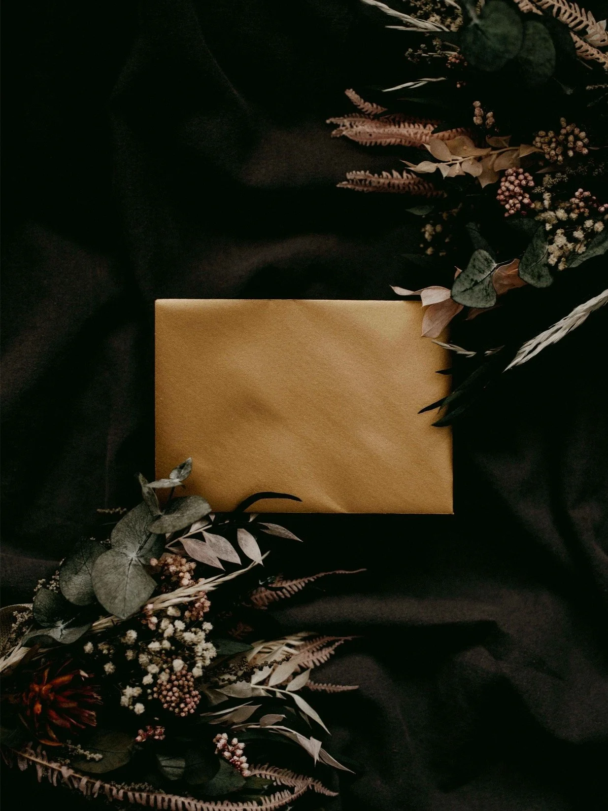 A flat lay photo of a yellow envelope surrounded by dark flowers and foliage, all placed on black fabric.