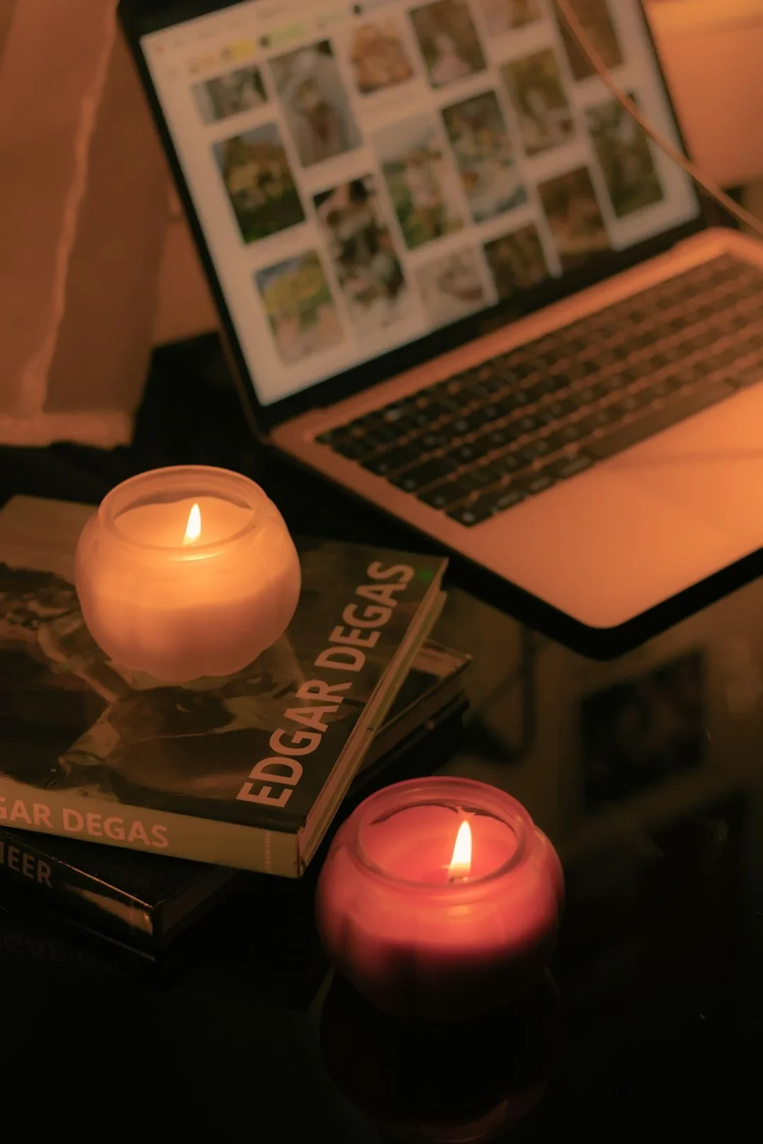 A laptop on a table with a photo gallery on its screen, surrounded by two lit candles and a couple of books, one titled 'Edgar Degas'.