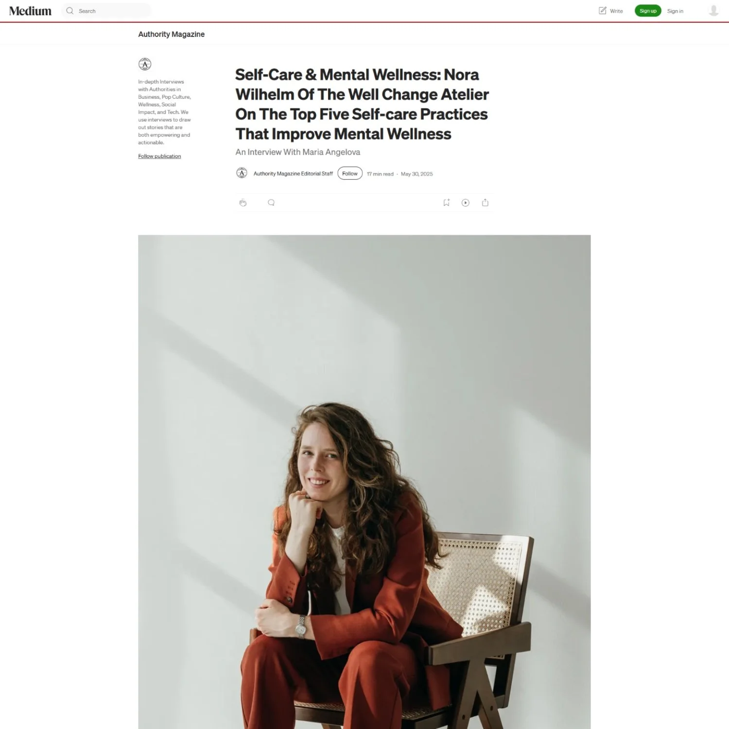 A woman with long curly hair sits on a wooden chair with a woven backrest, wearing a rust-colored suit, smiling softly and resting her chin on her right hand, against a plain light-colored wall with shadows.