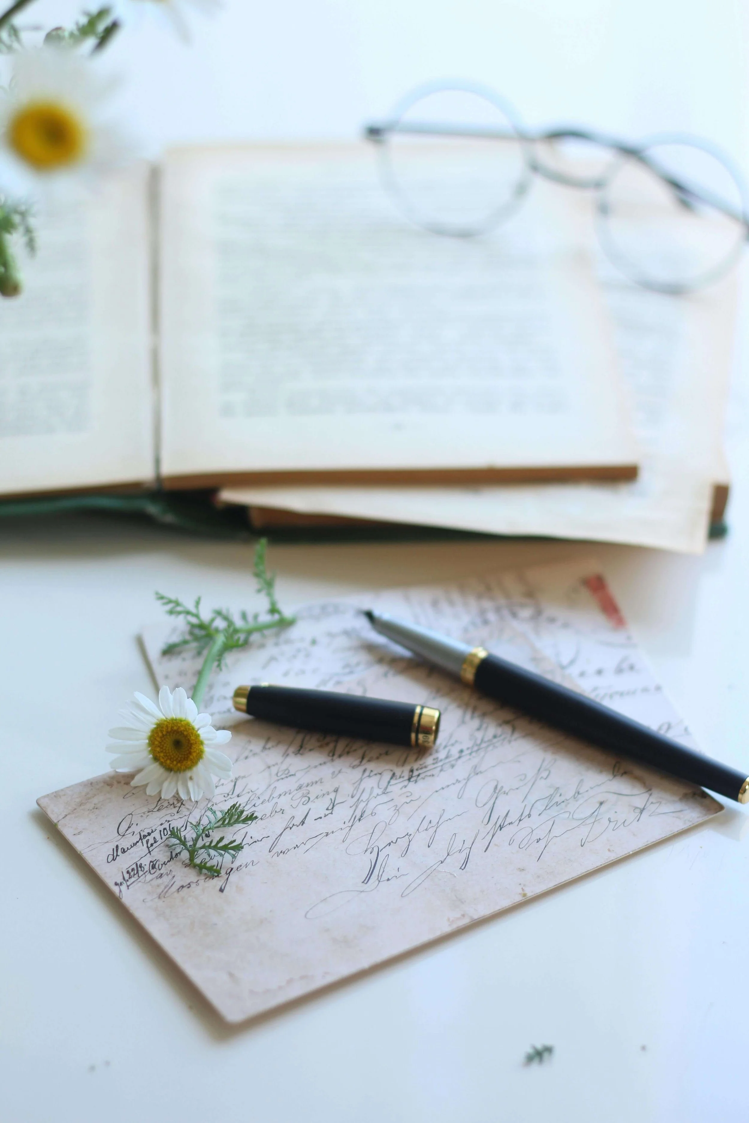 Open book with round glasses, a flower, pen, and handwritten letter on a white surface.