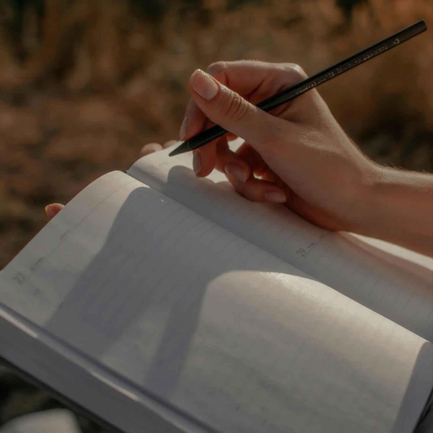Close-up of a person's hand writing in a notebook outdoors with a black pen, with sunlight casting shadows on the pages.